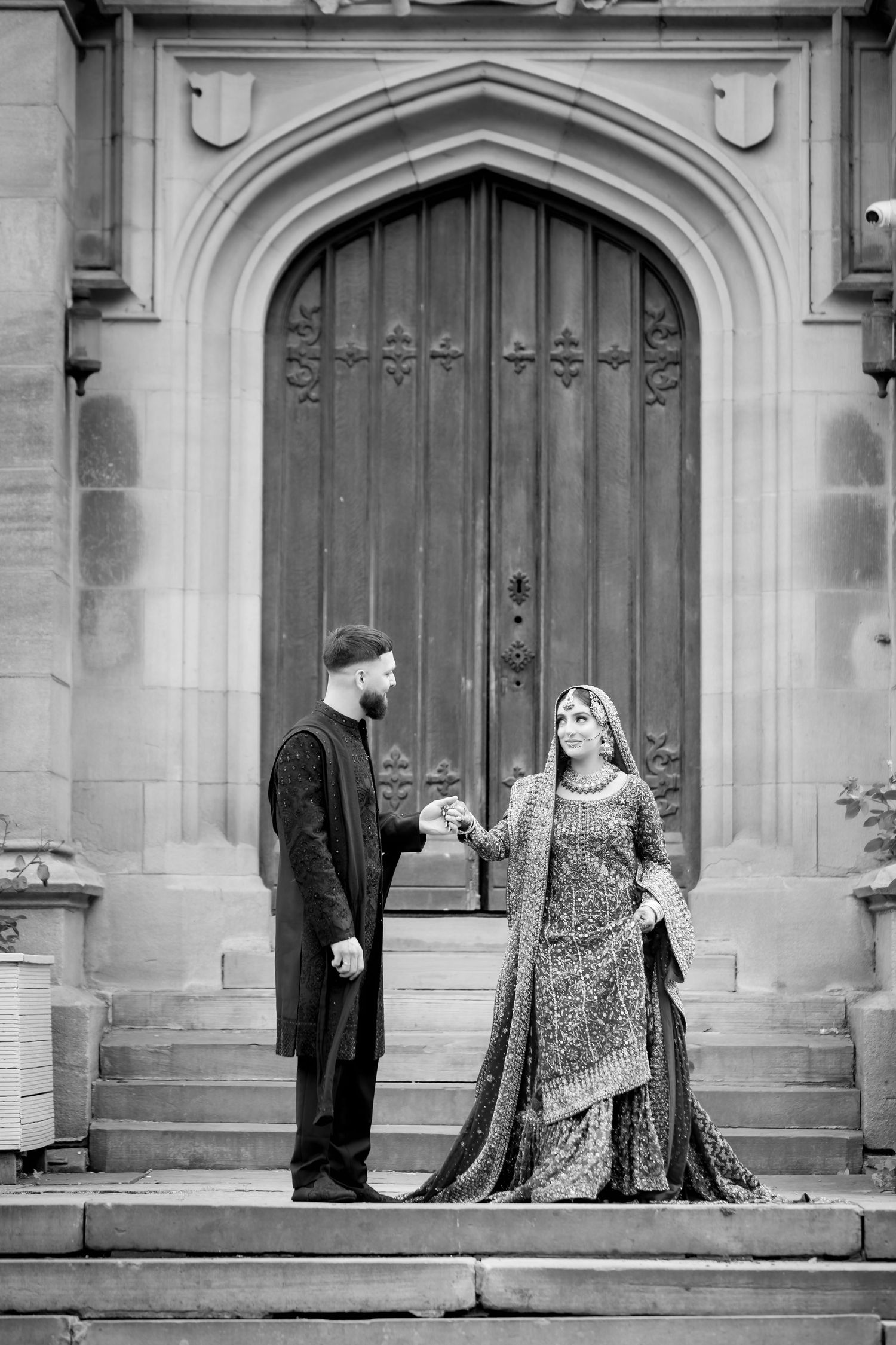 Black and white photo of South Asian couple in traditional attire holding hands on steps before ornate arched wooden door.