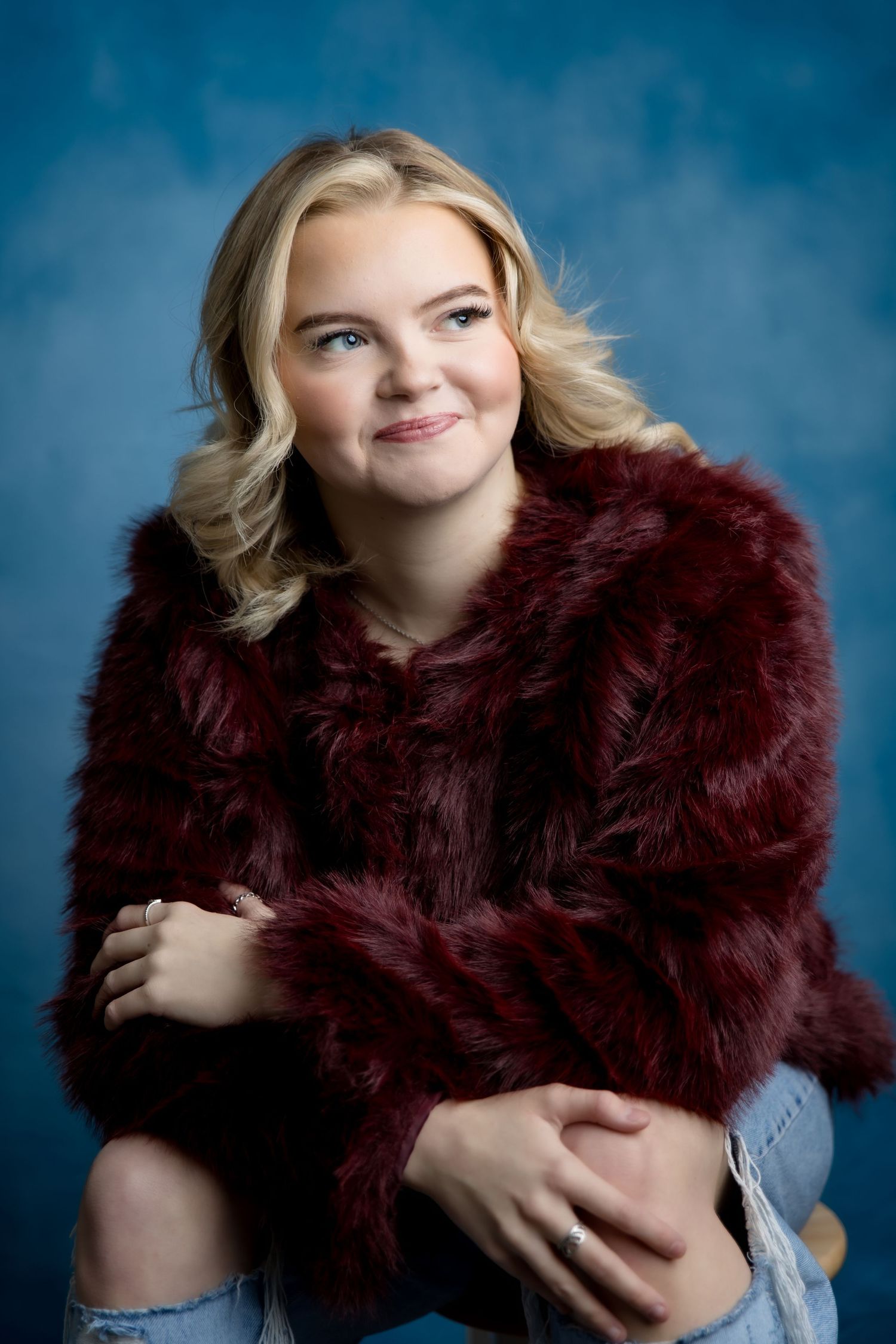 Young blonde woman in burgundy faux fur jacket and ripped jeans poses against blue studio backdrop.