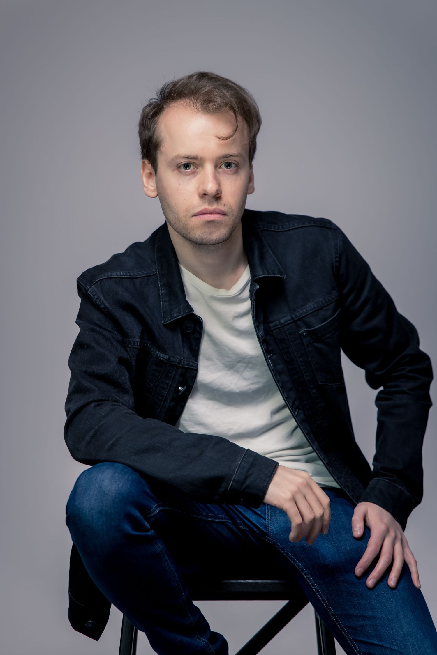 Young man in black denim jacket and jeans sitting on stool against gray background in studio portrait.