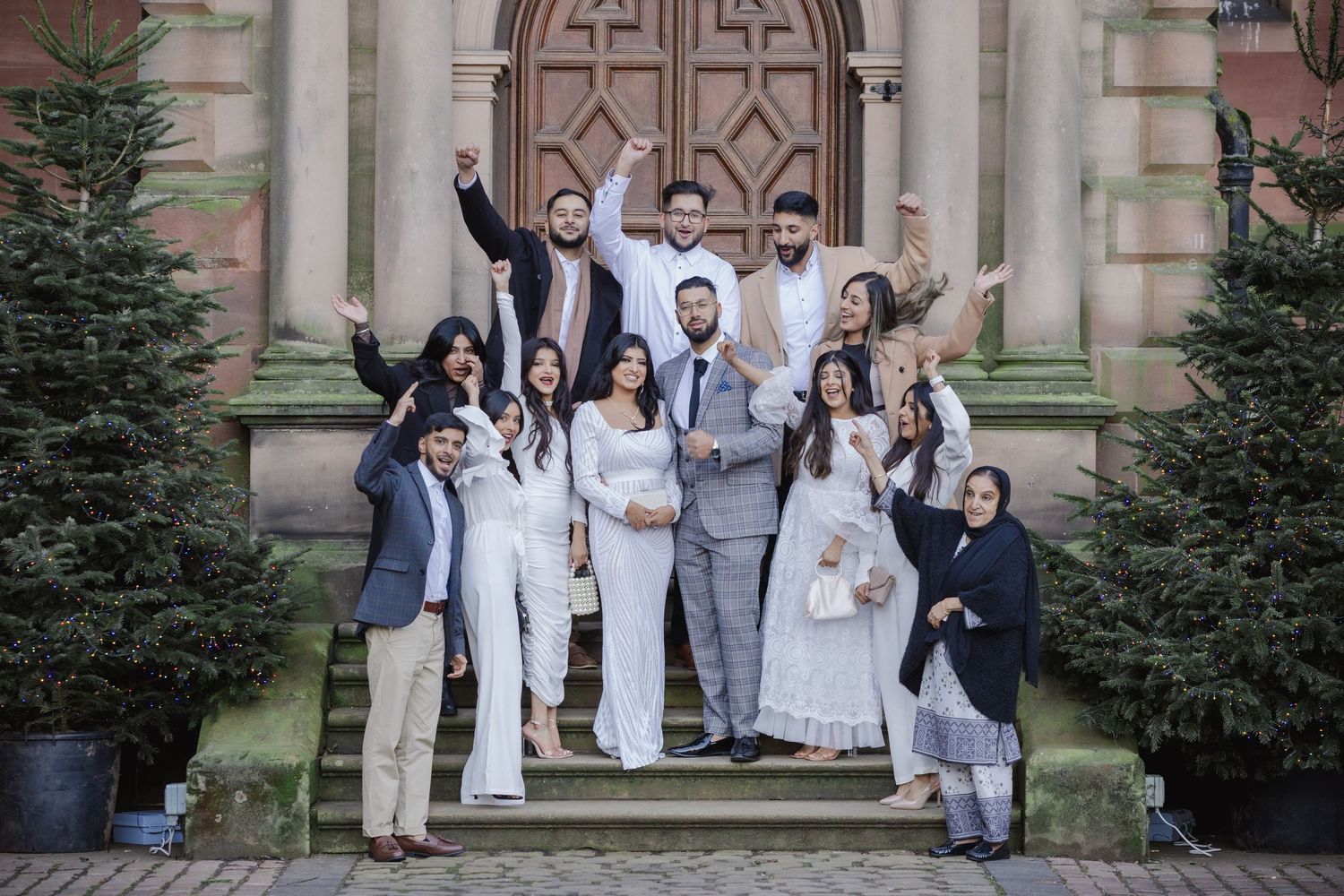 Large family group celebrating on church steps, dressed in elegant white and gray outfits, raising hands joyfully.