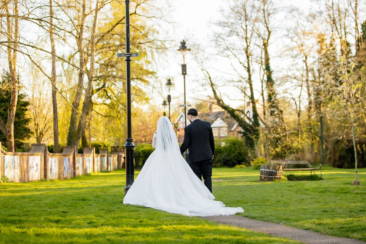 A bride and groom walk hand in hand through a lush green park, with the bride's white gown trailing behind them.