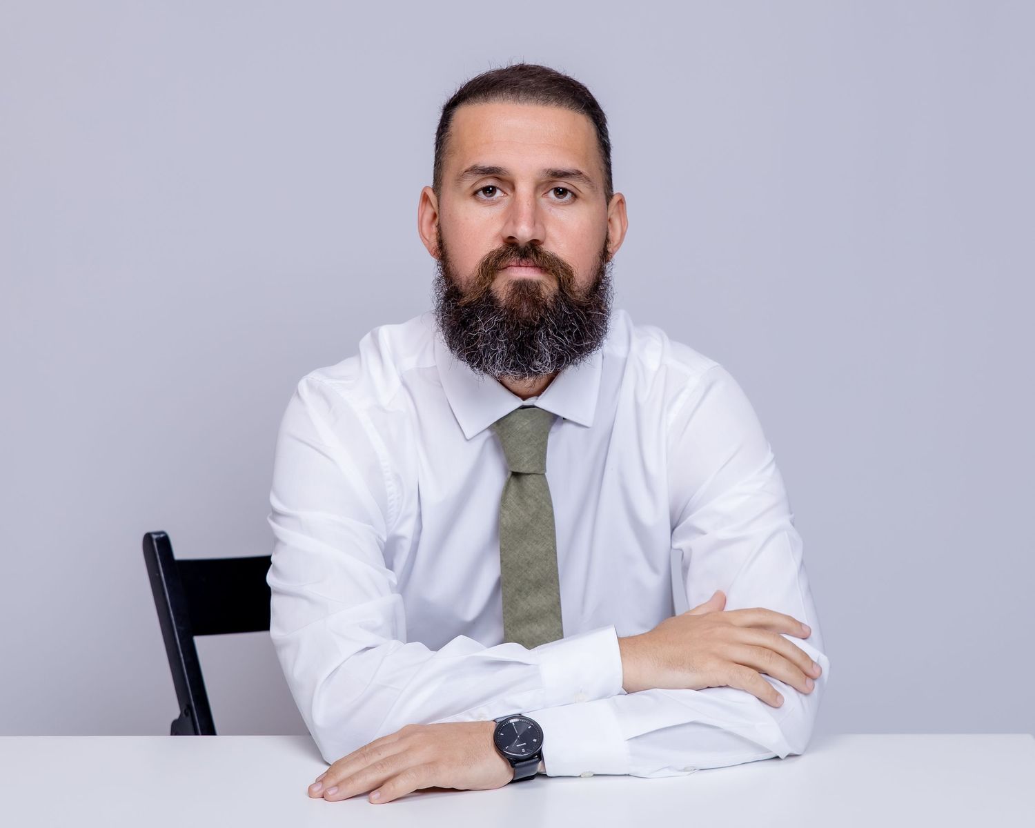 Professional bearded man in white shirt and olive tie sits with arms crossed at white desk.