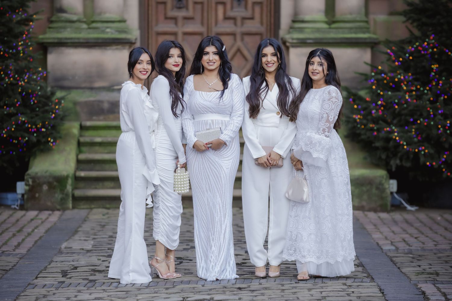 Five women in elegant white outfits posing together outside a historic building decorated for Christmas.