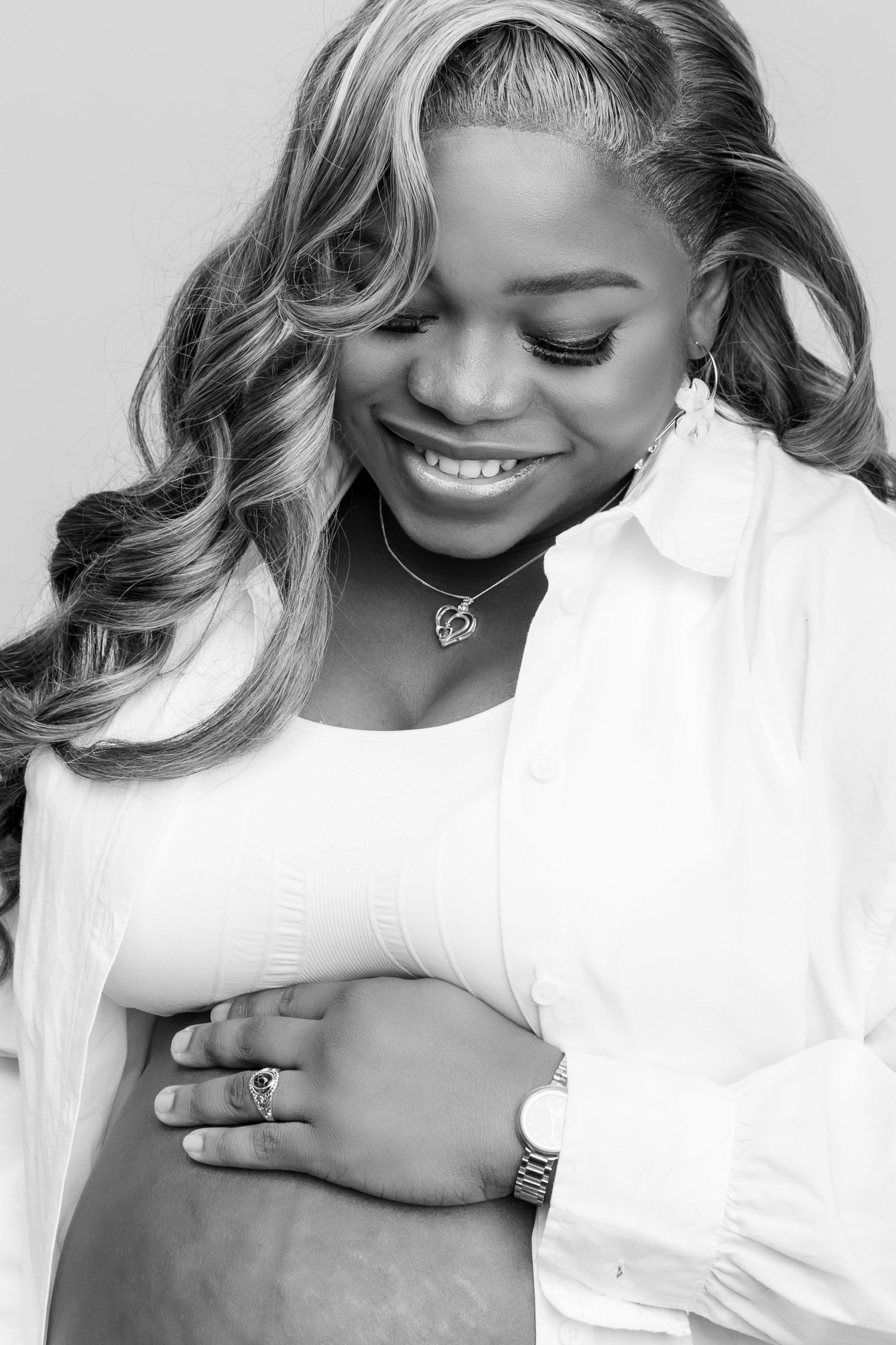 Black and white portrait of a smiling pregnant woman with long wavy hair, wearing a white shirt and necklace.