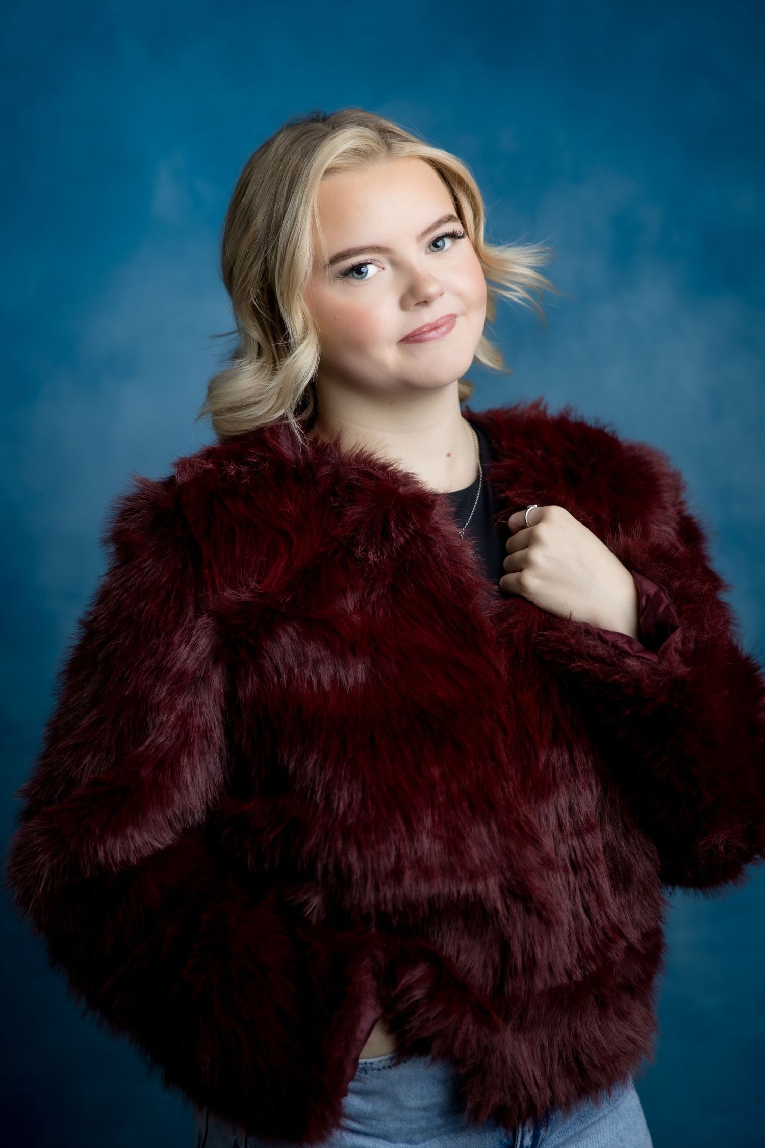 Young blonde woman posing in a dark red faux fur jacket against a blue studio background.