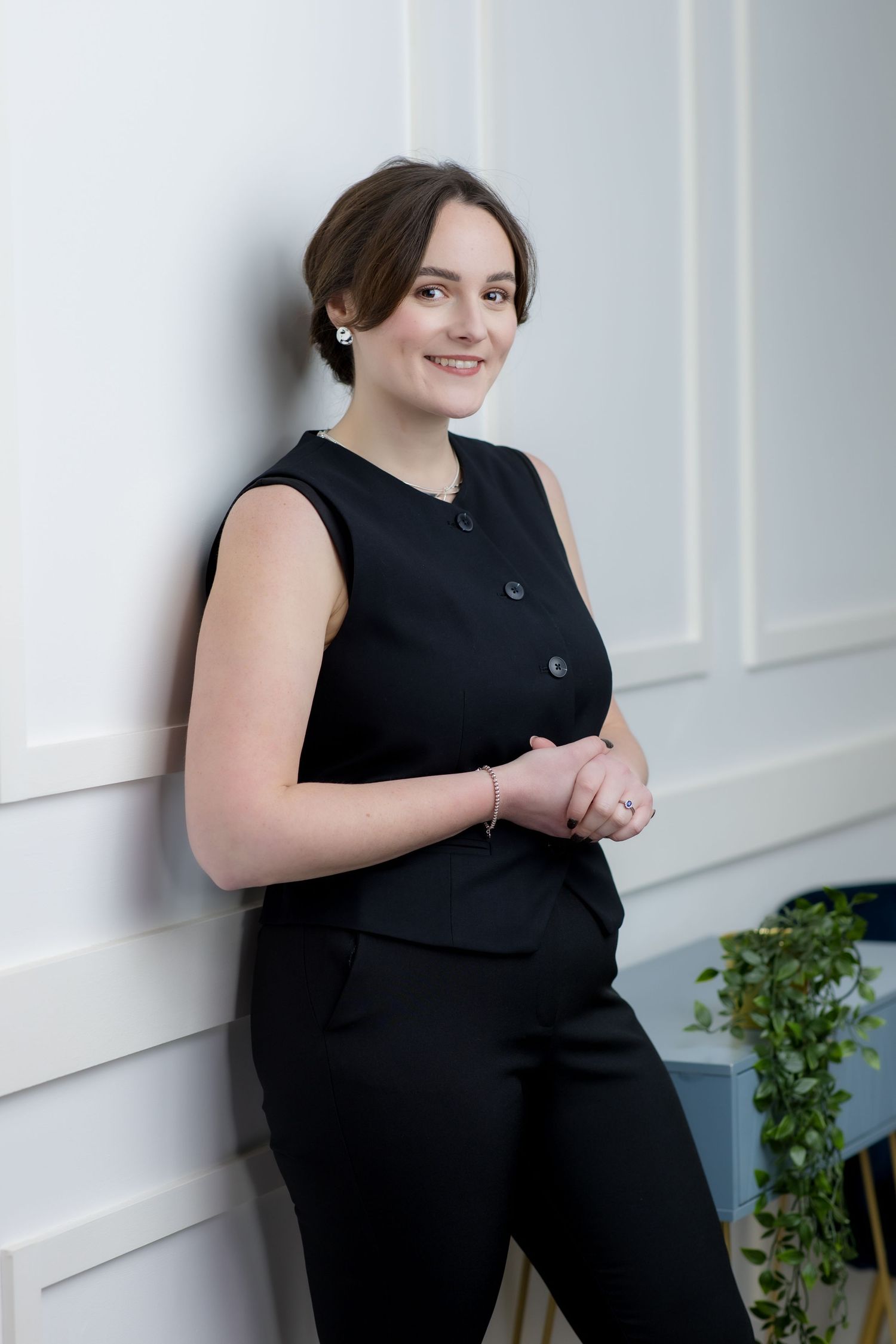 Young woman in black sleeveless pantsuit smiling and leaning against a white wall in a professional portrait.