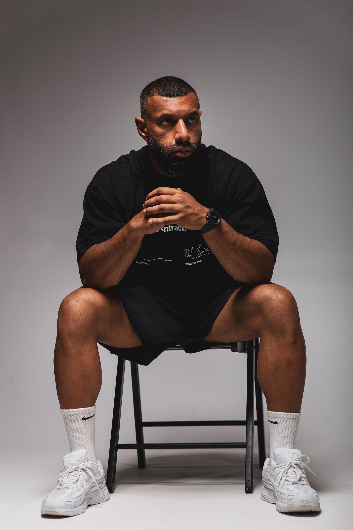 Muscular man in black outfit sitting on chair wearing Nike socks and white sneakers against gray background.