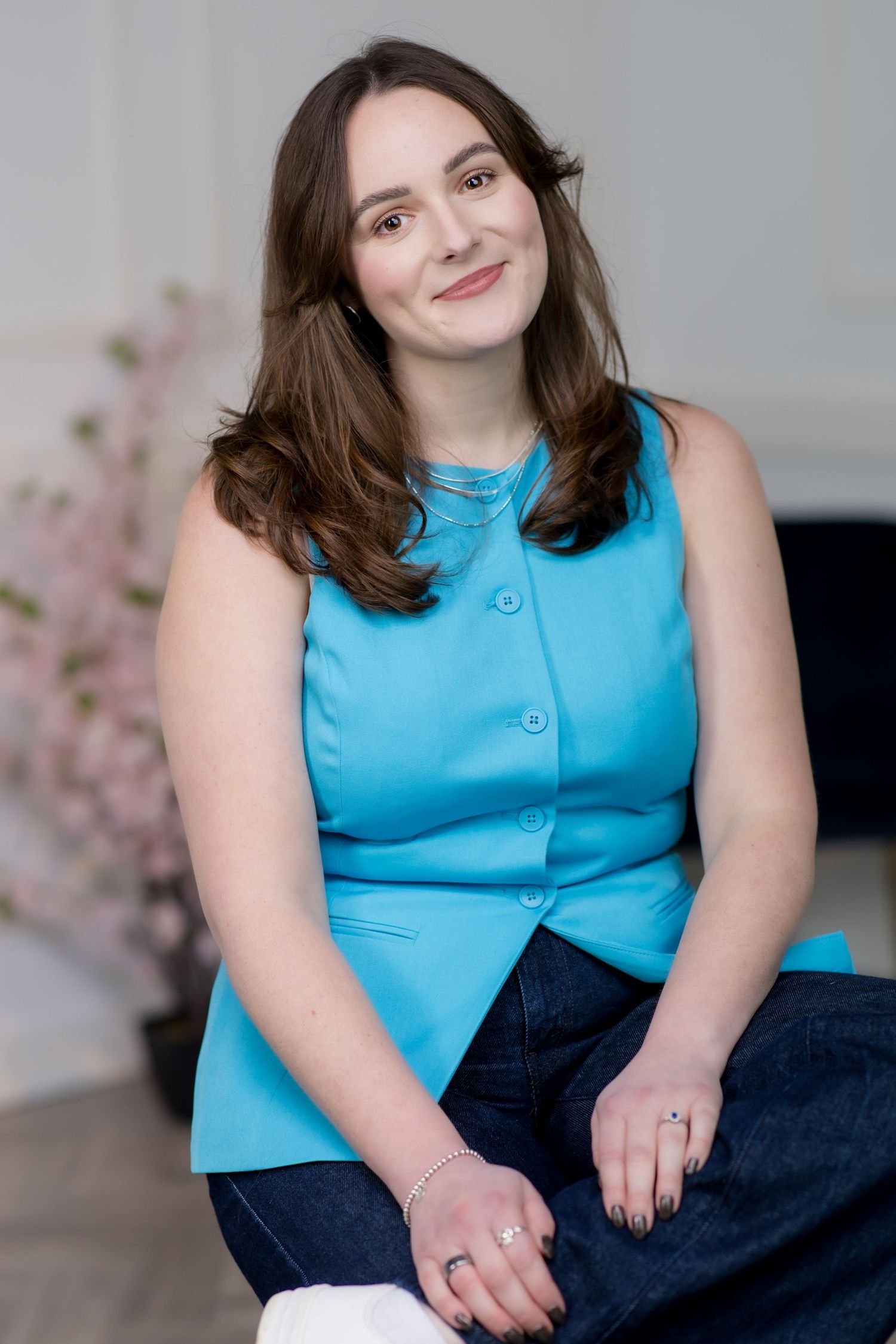 Young woman in a teal sleeveless top and dark jeans sits smiling in a bright room with pink flowers.