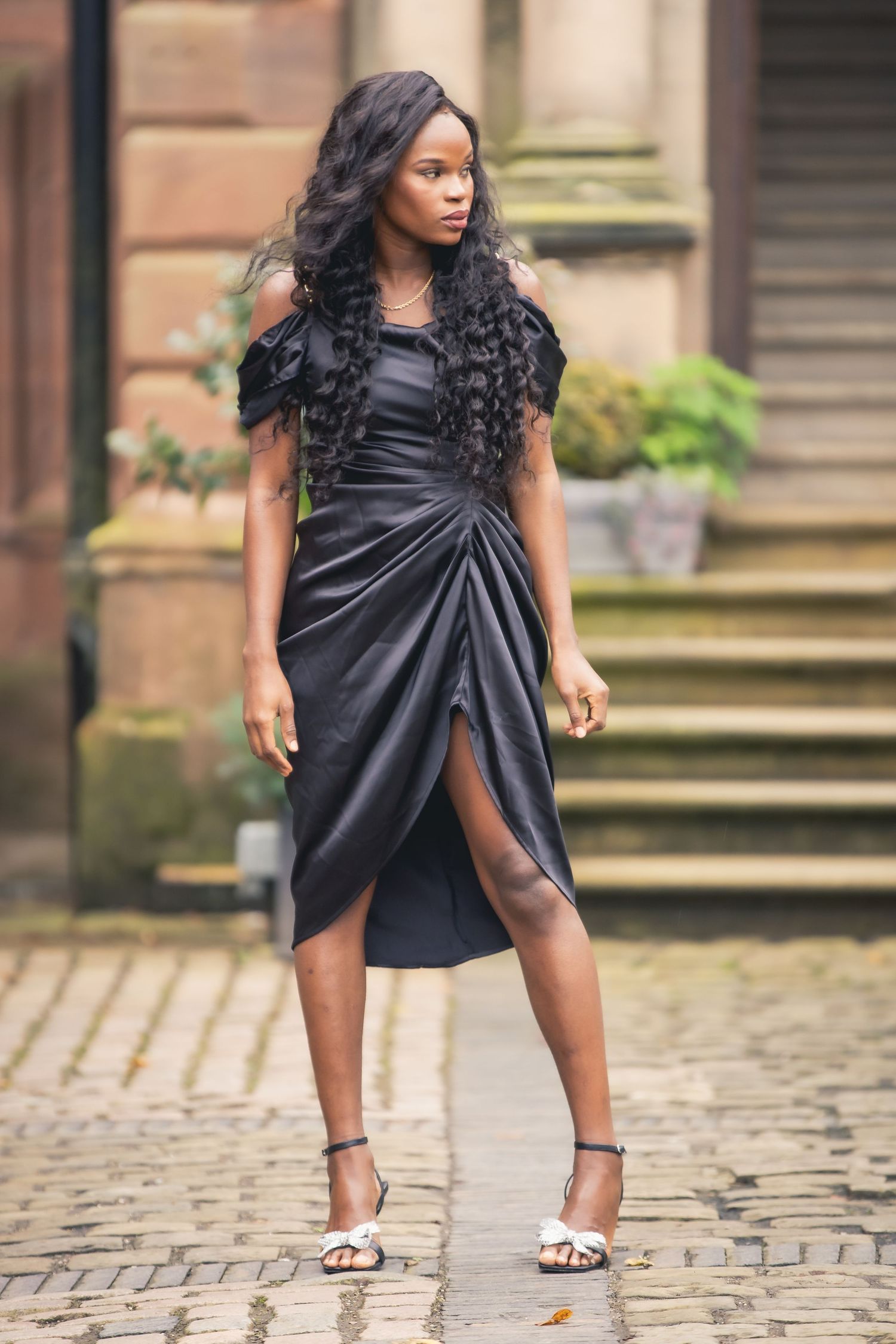 Elegant Black woman in a black satin ruffle dress and silver heels posing on cobblestone street.
