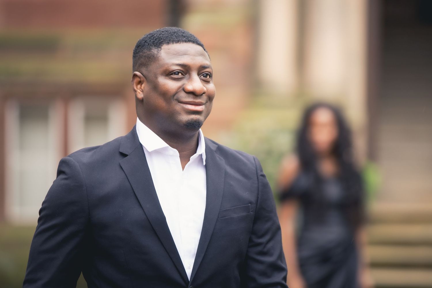 A well-dressed Black man in a dark suit smiles confidently outdoors, with a woman standing in the background.