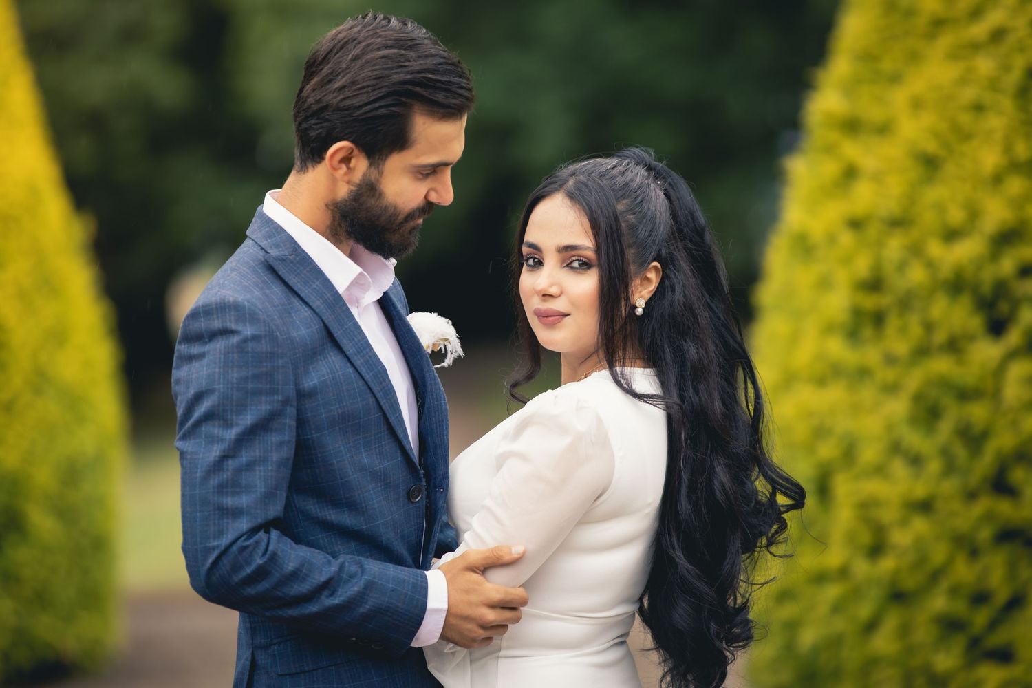 Elegant couple in wedding attire embracing in a garden, groom in blue suit, bride in white dress with long dark hair.