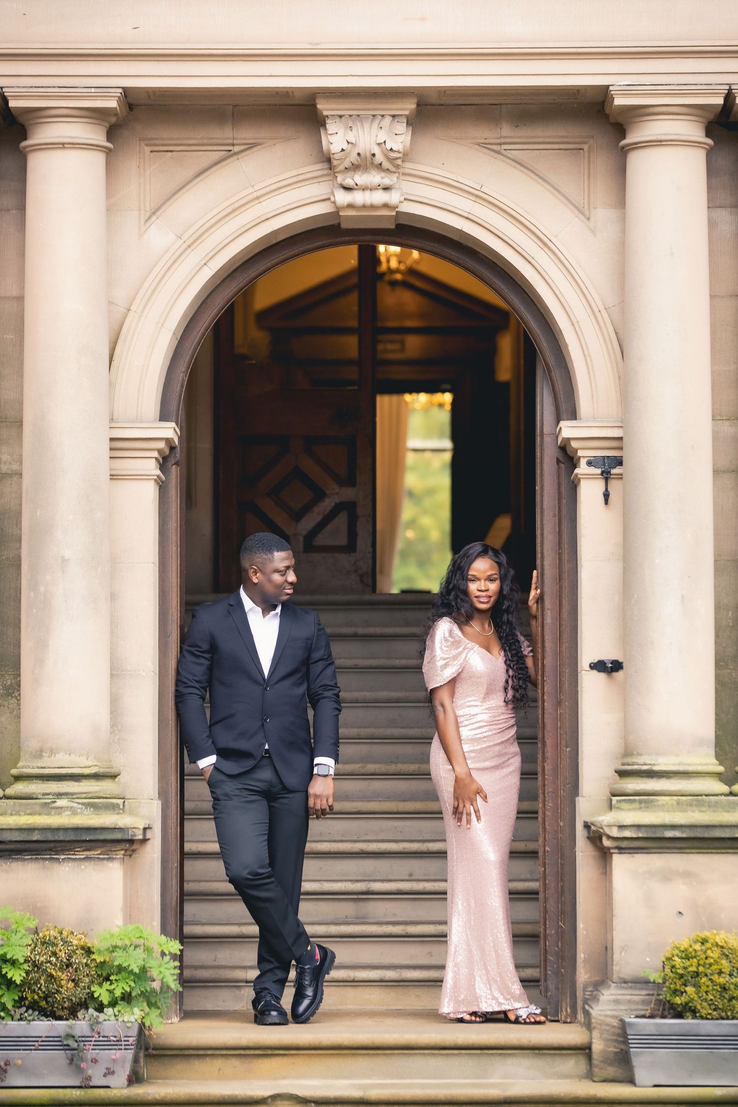 Elegant couple in formal attire standing in grand arched stone doorway on steps.