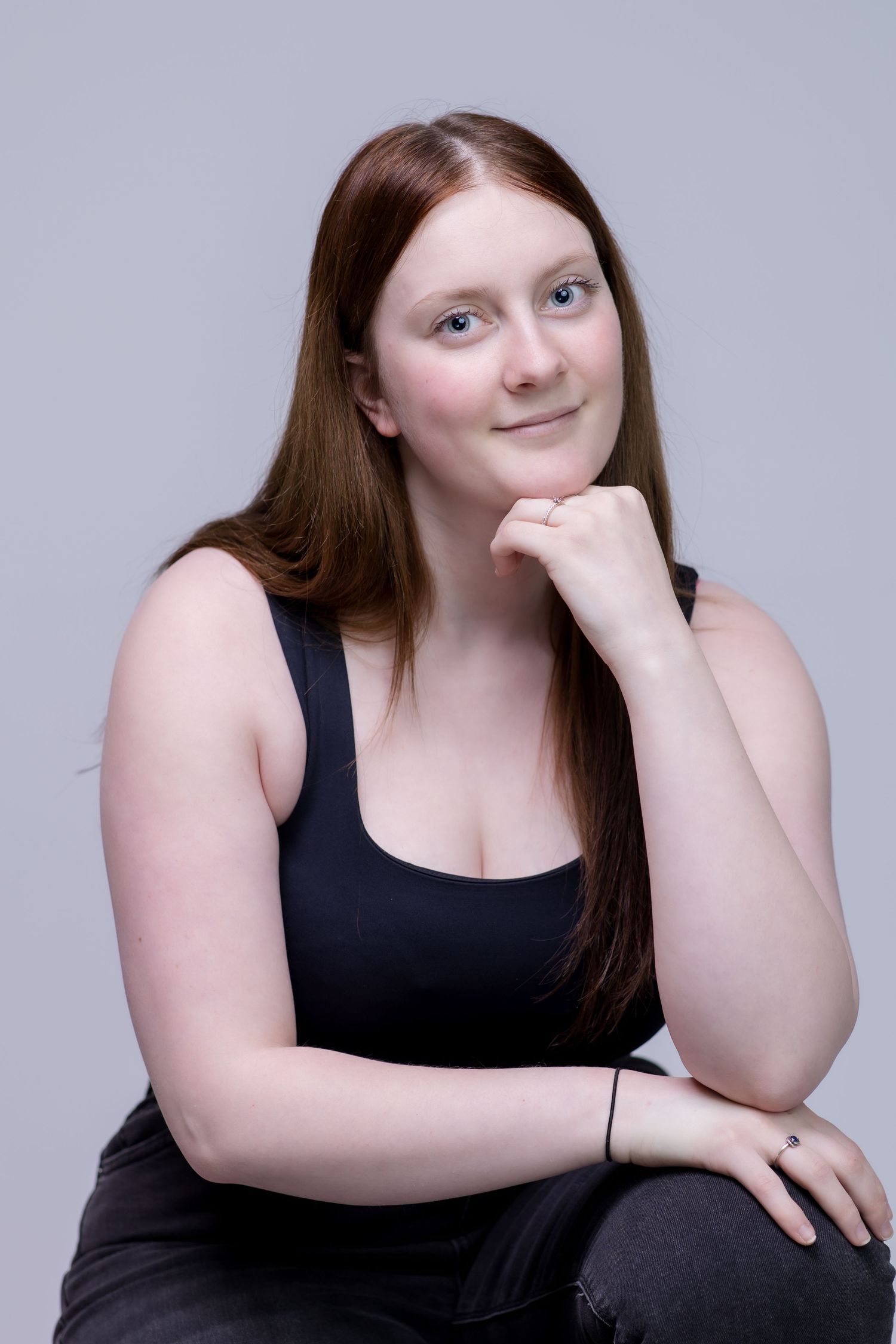 Young woman with long red hair in black tank top poses thoughtfully with hand on chin against gray background.