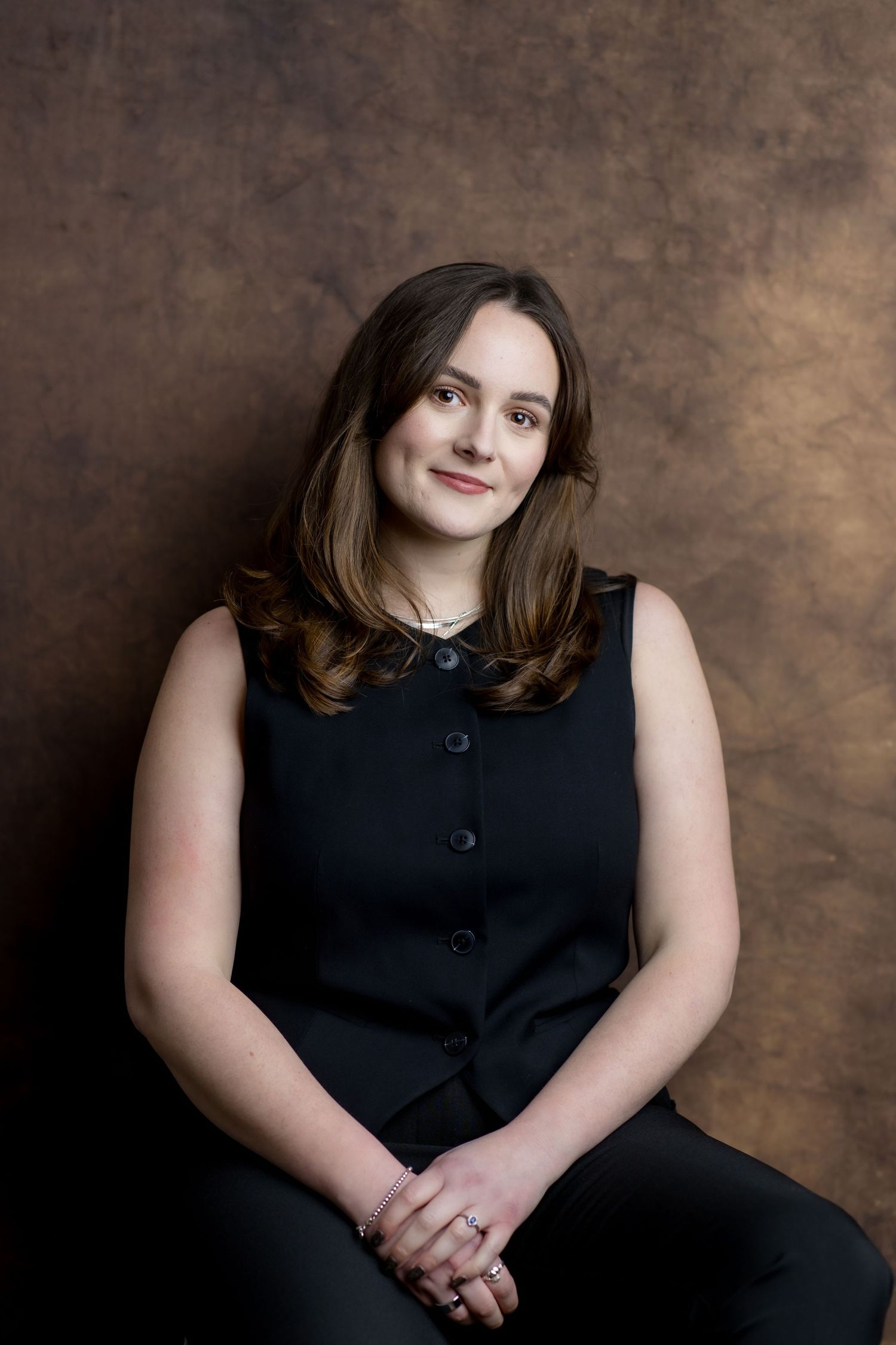 Professional woman in black sleeveless dress seated, smiling confidently against a brown studio backdrop.