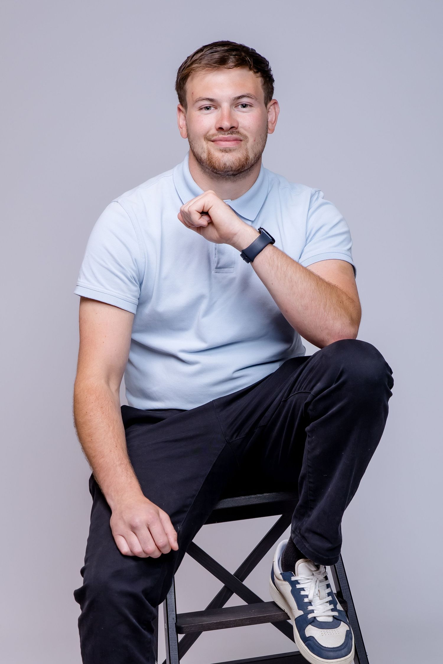 Young man in light blue polo shirt sitting on stool with crossed legs, wearing smartwatch, smiling at camera.
