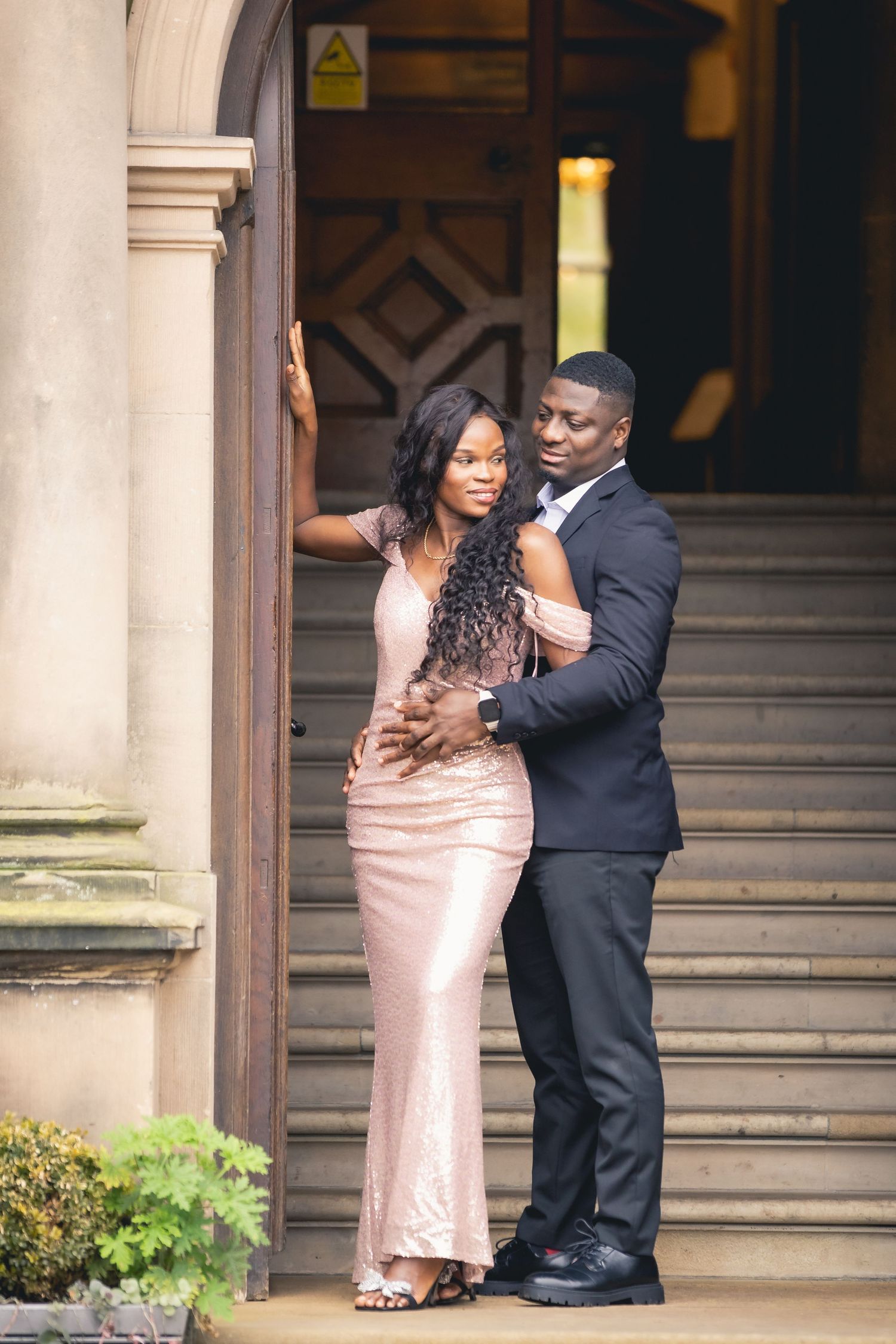 Elegant couple posing at grand building entrance, woman in rose gold gown, man in dark suit, on stone steps.