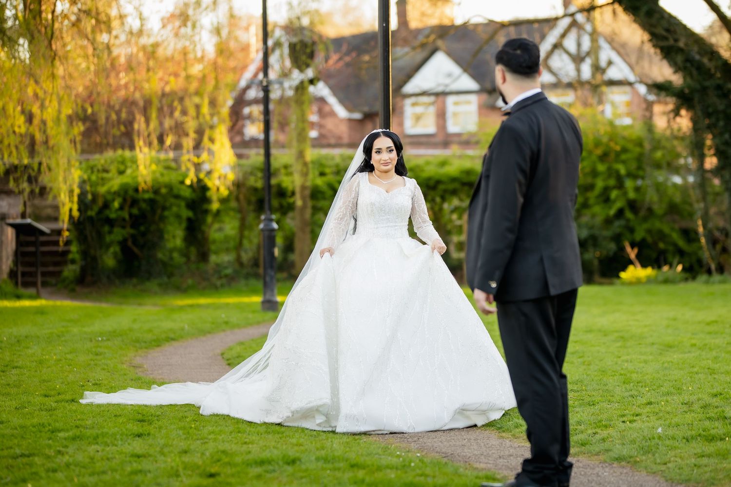 Bride in elegant white ball gown with veil faces groom in black suit on garden path at English countryside wedding venue.