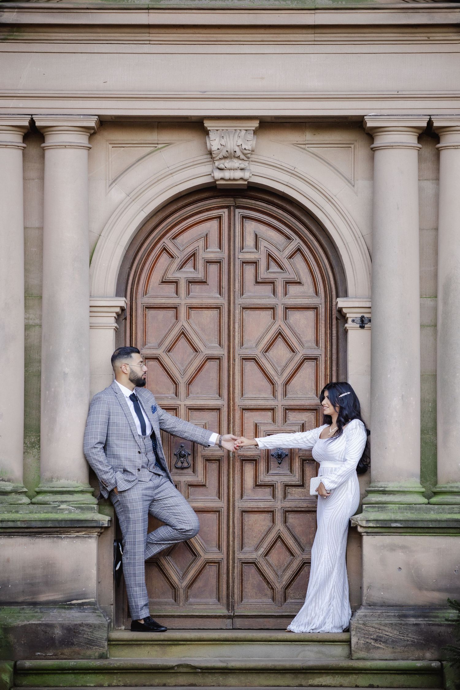 Couple in wedding attire holding hands before ornate wooden arched door of historic stone building.