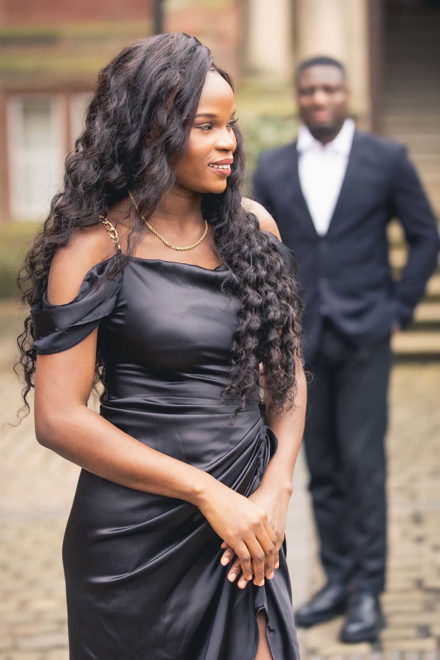Stylish Black woman in black satin dress smiling outdoors with man in suit standing behind her.