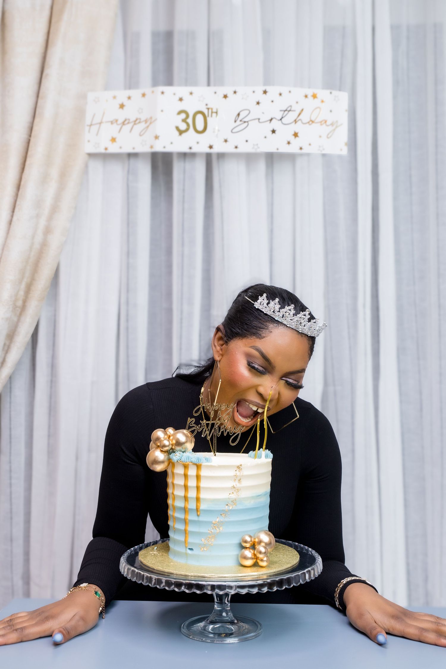 Woman in black dress and tiara laughing beside elegant blue and gold 30th birthday cake at celebration.