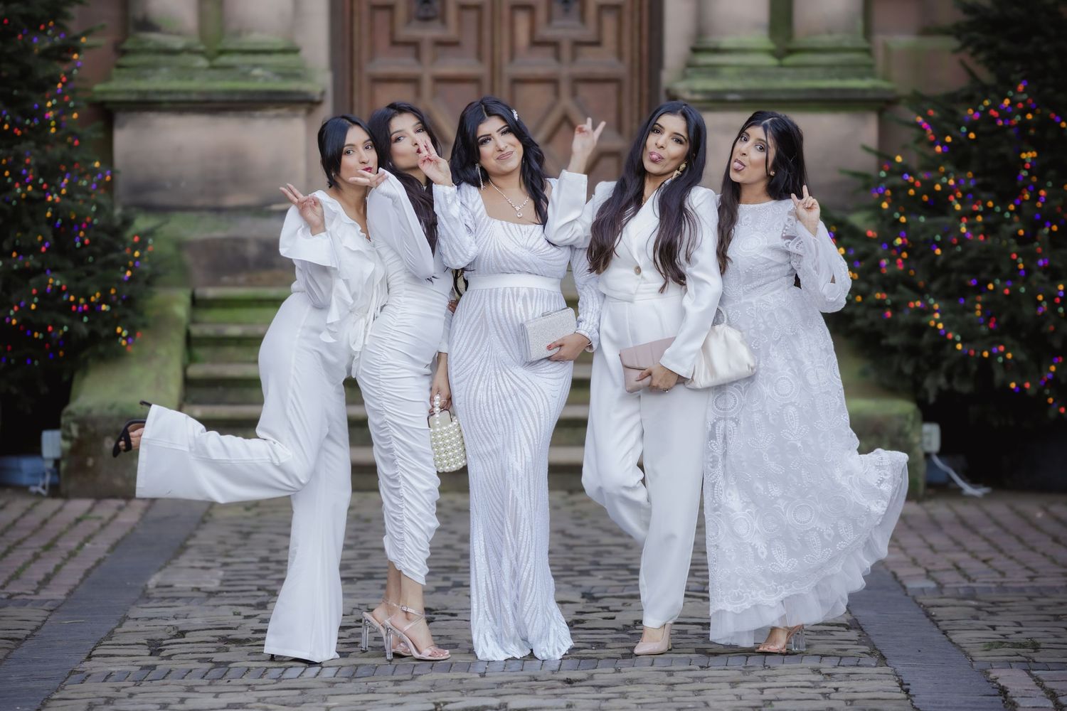 Five women in elegant white outfits posing together outdoors near Christmas trees and stone architecture.