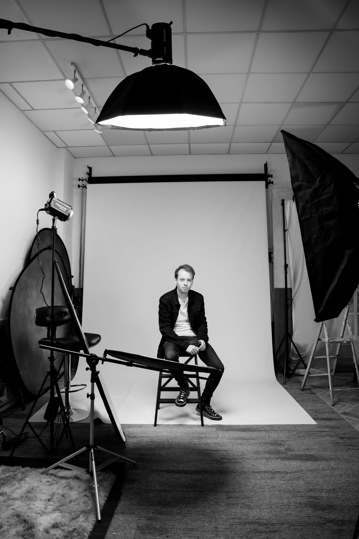 Black and white photo of a man sitting on a stool in a professional photography studio with lighting equipment.