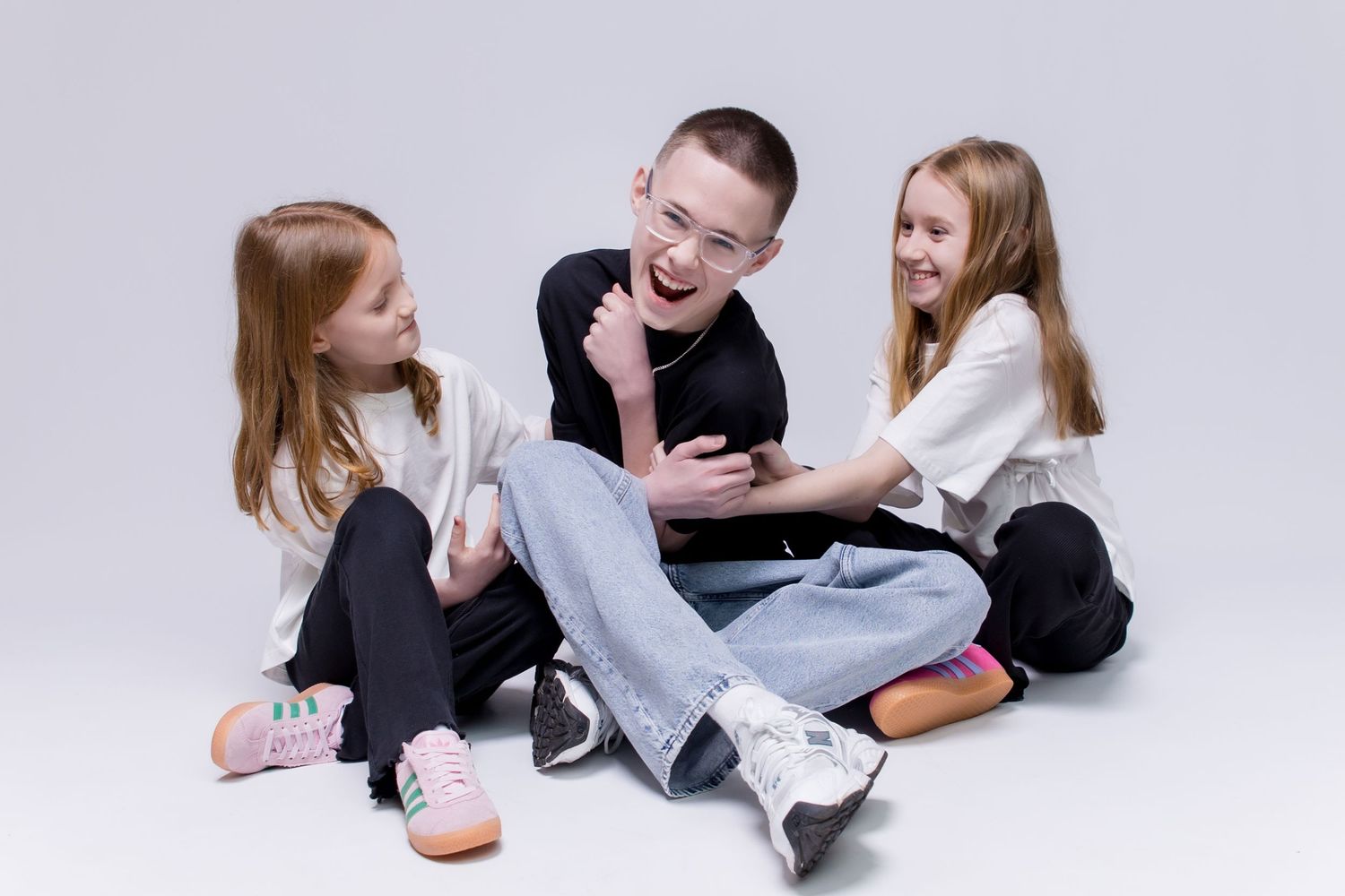 Three children laughing and playing together while sitting on a white studio floor in casual clothing.