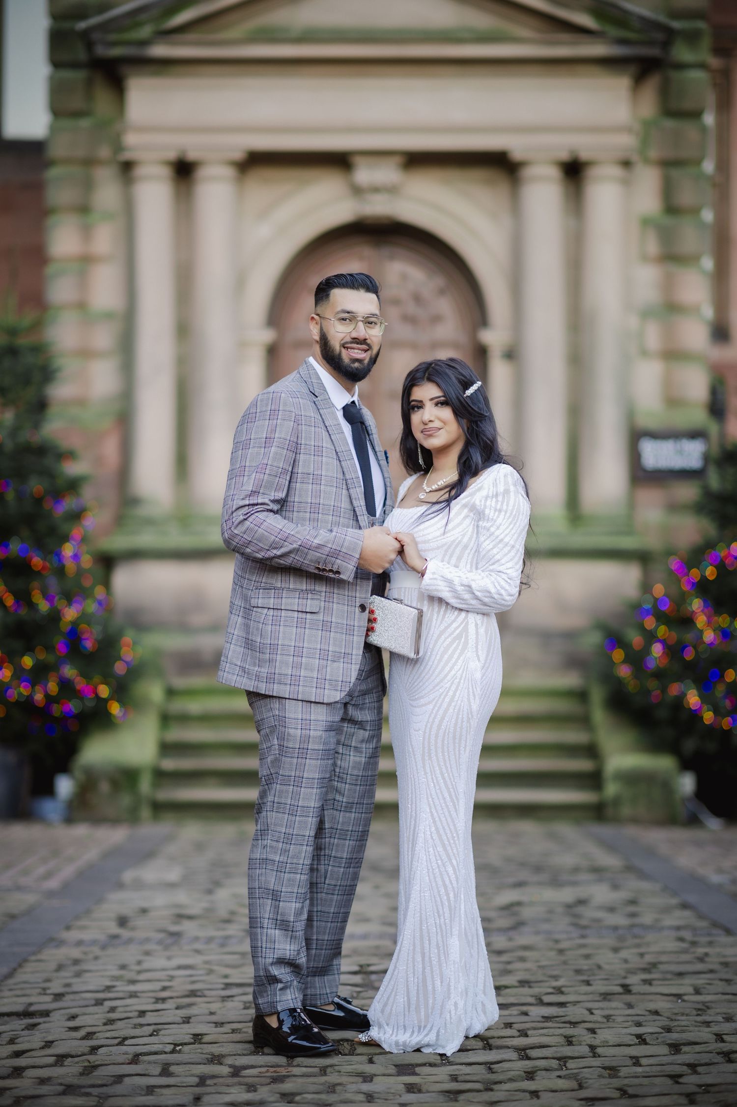 Elegant couple posing in front of a stone building, man in plaid suit and woman in white sequin gown.