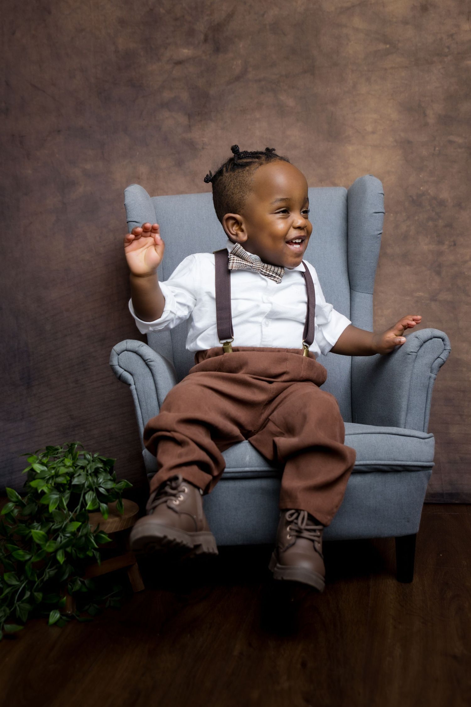 Adorable toddler boy in suspenders, bow tie, and brown pants sitting in a blue armchair, laughing joyfully.
