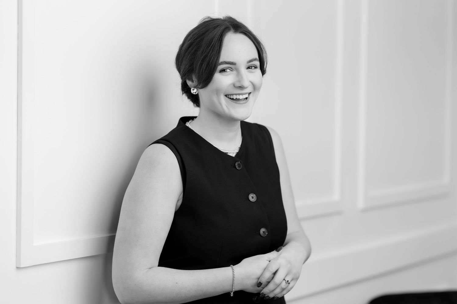 Professional black and white portrait of a smiling young woman in a sleeveless black top leaning against a white wall.