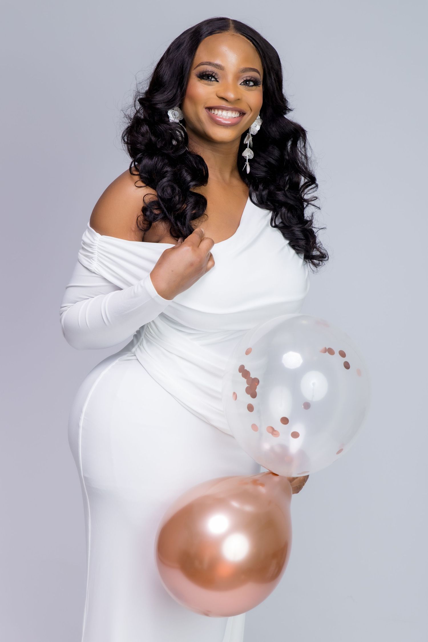 Smiling pregnant woman in white off-shoulder dress holding rose gold and clear confetti balloons on gray background.