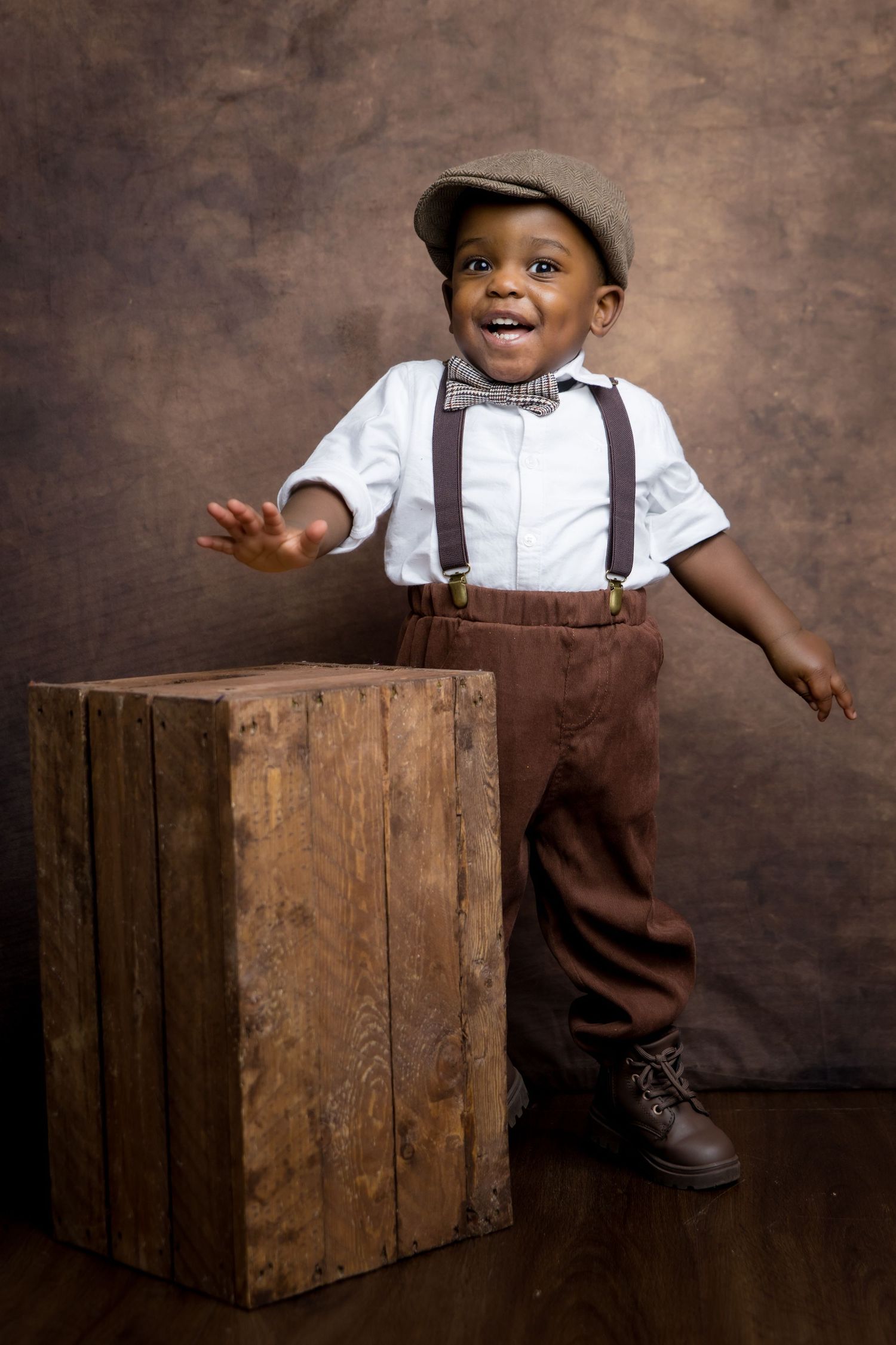Adorable young Black boy in vintage outfit with suspenders, bow tie, flat cap, and brown pants posing by wooden crate.