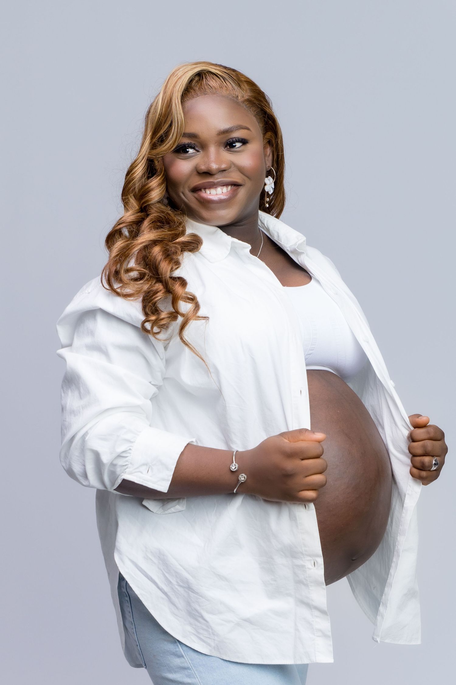 Smiling pregnant Black woman with curly blonde hair, wearing white shirt and jeans, holding open shirt to show baby bump.