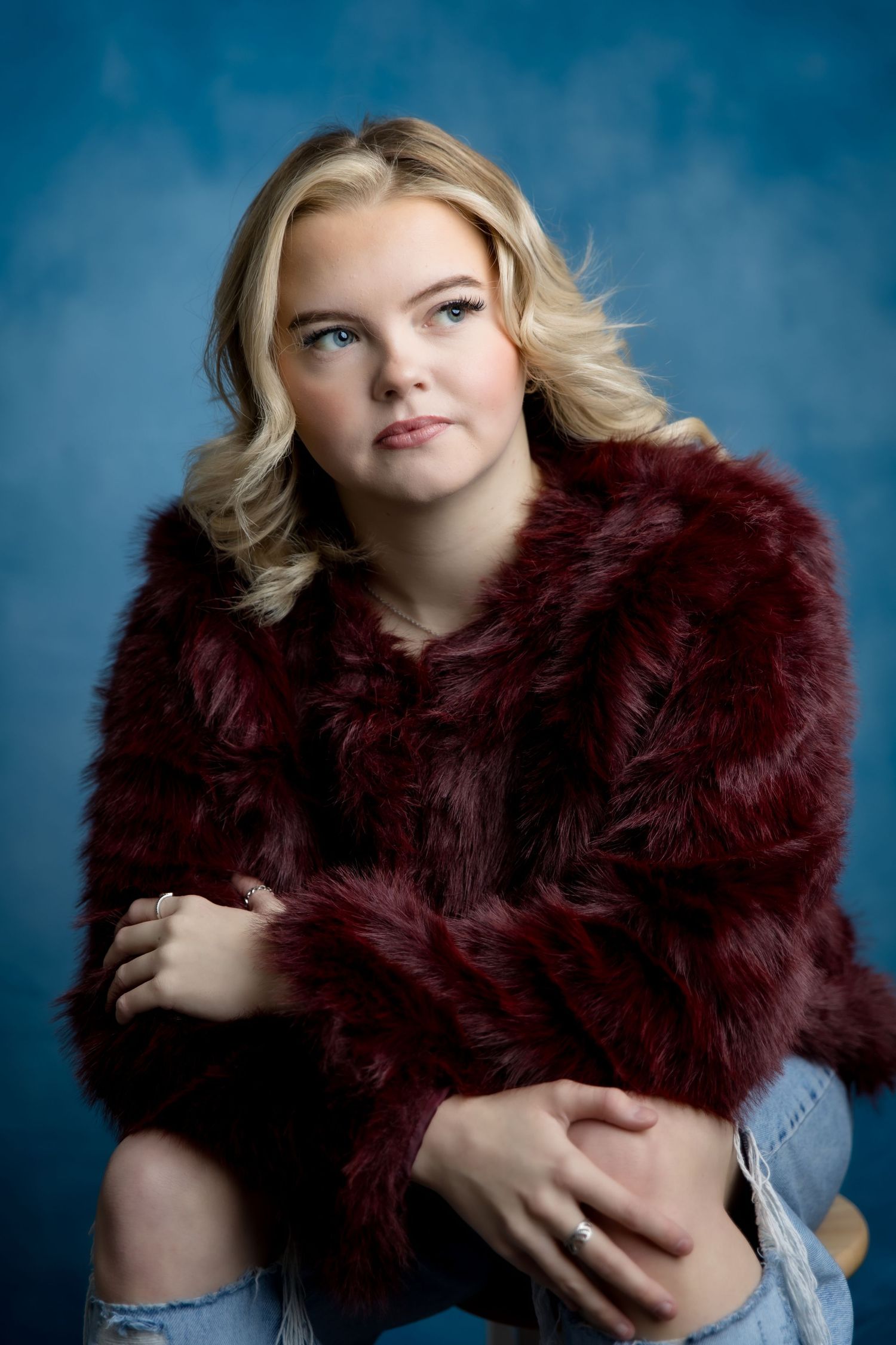Young blonde woman in burgundy faux fur coat and ripped jeans poses against blue studio background.