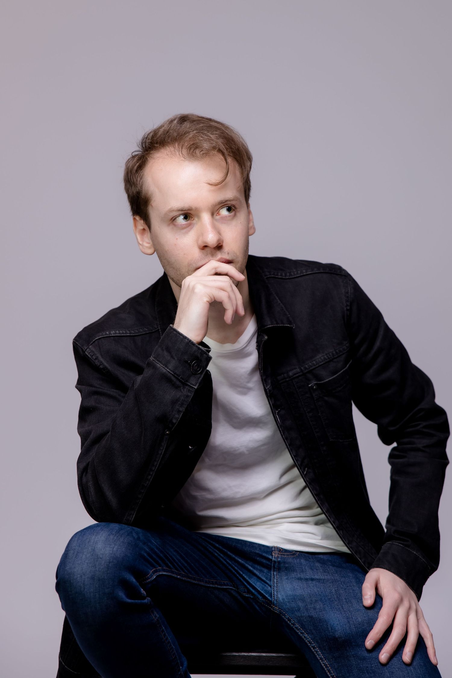 Thoughtful young man in black denim jacket and jeans sitting and resting chin on hand against gray background.
