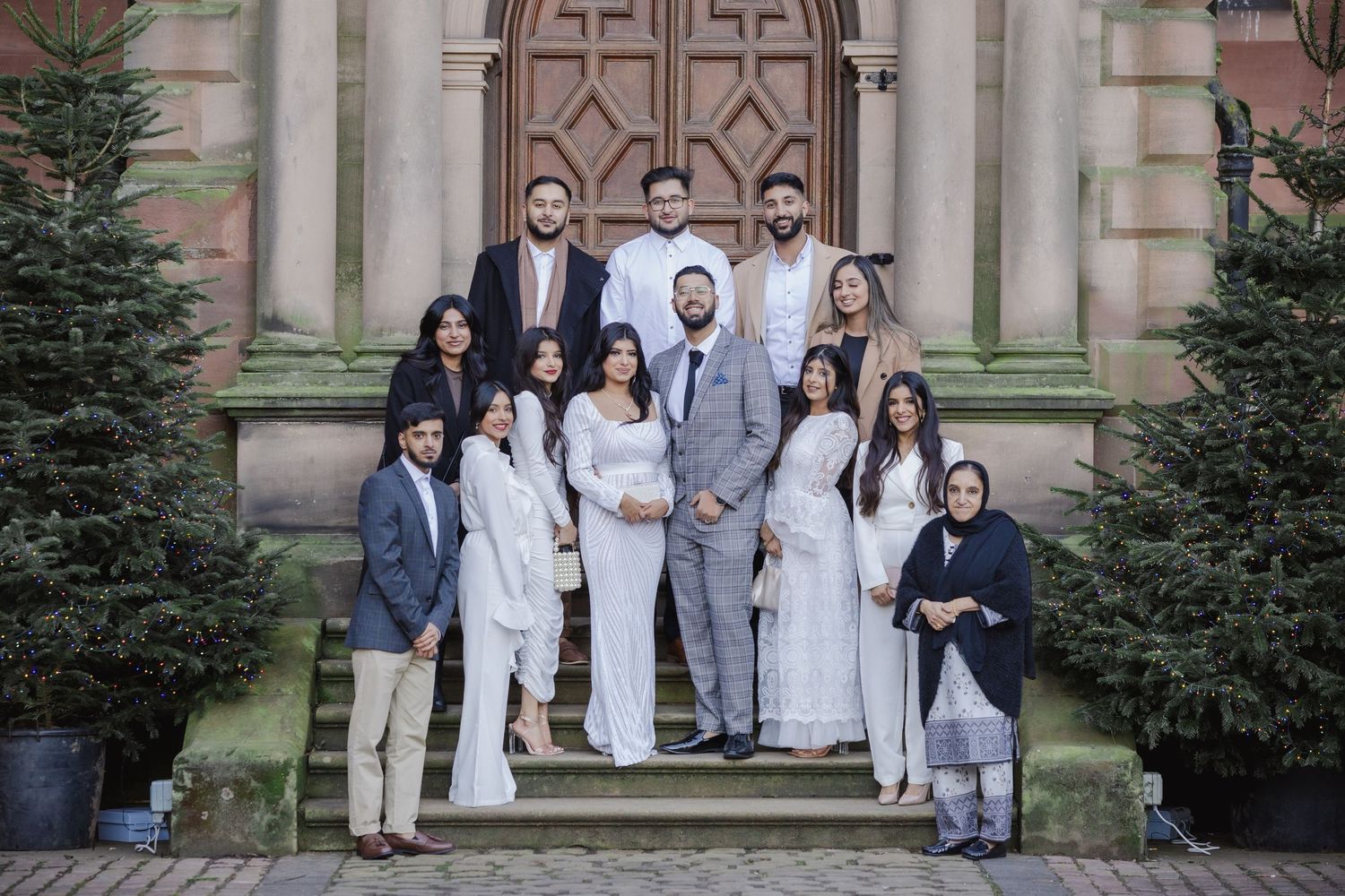 Large South Asian family group portrait in white outfits on church steps, flanked by Christmas trees.