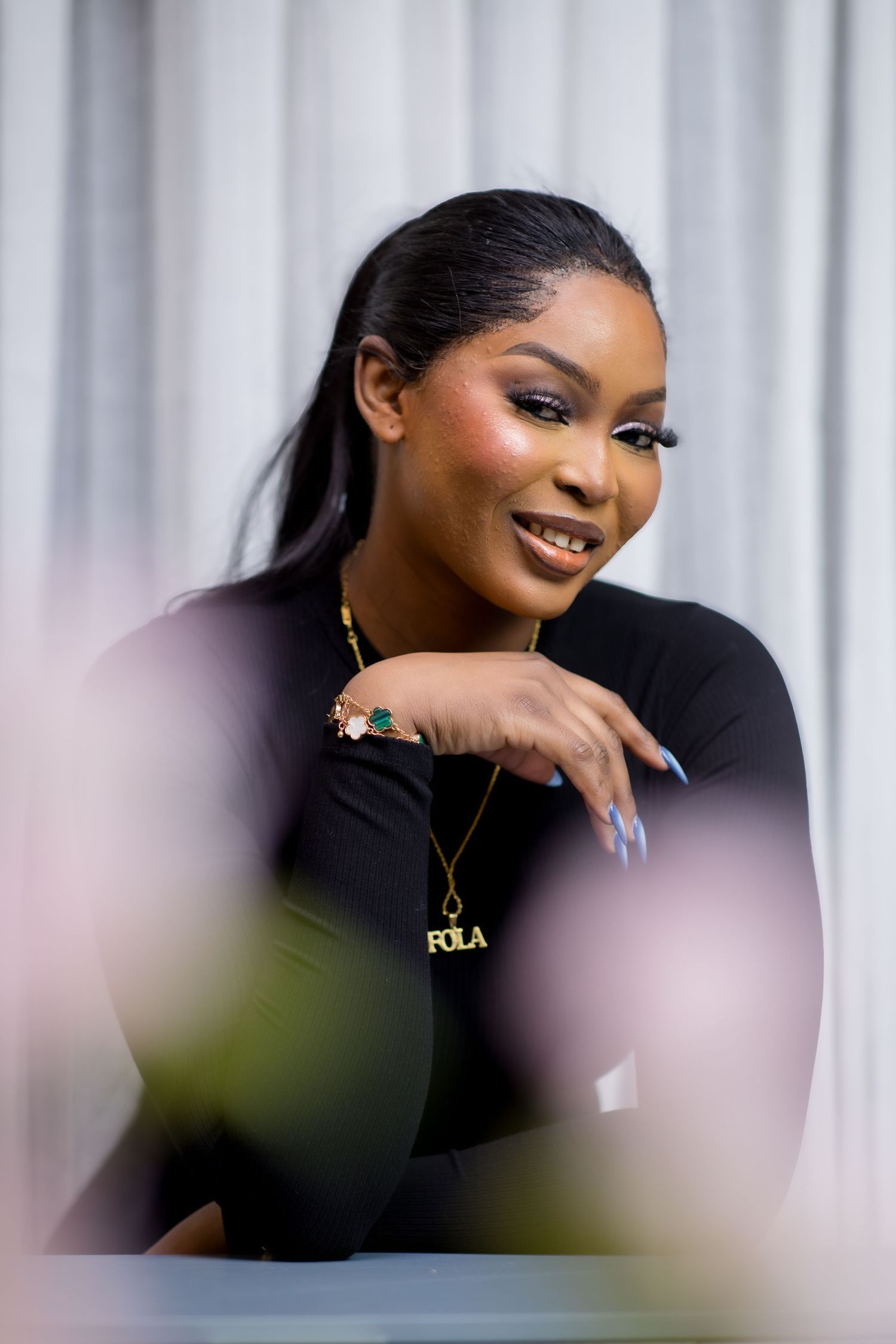 A young woman wearing a black outfit and gold FOLA nameplate necklace smiles gracefully in a portrait photo.