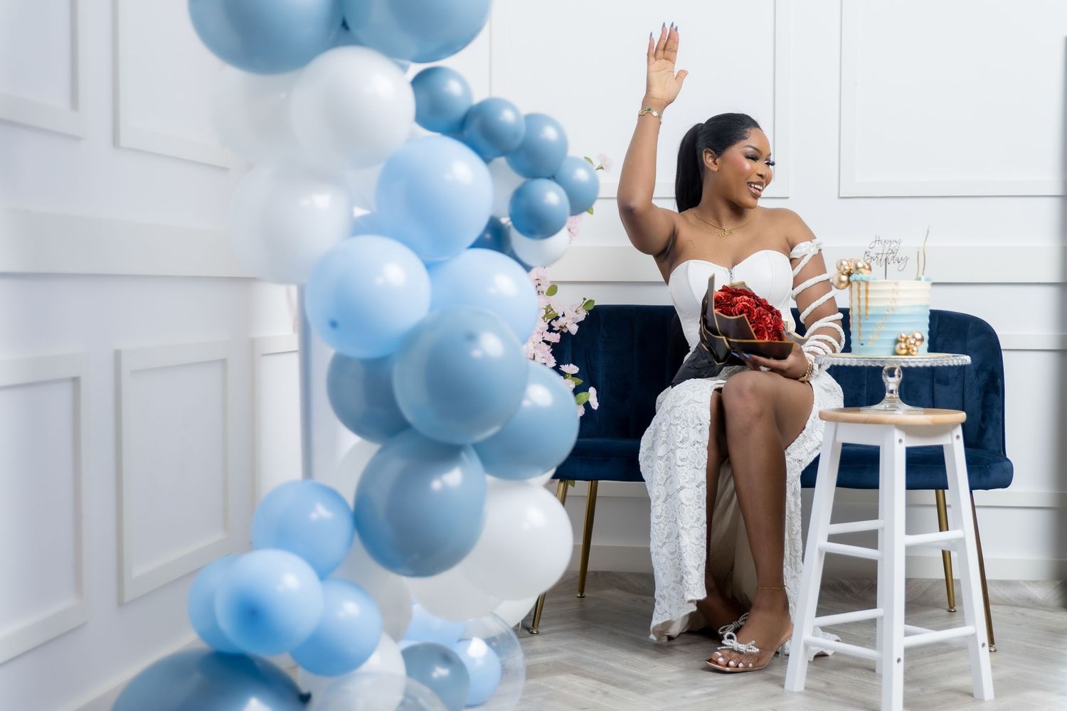 Woman in white dress waving at a baby shower, surrounded by blue and white balloons and gifts.