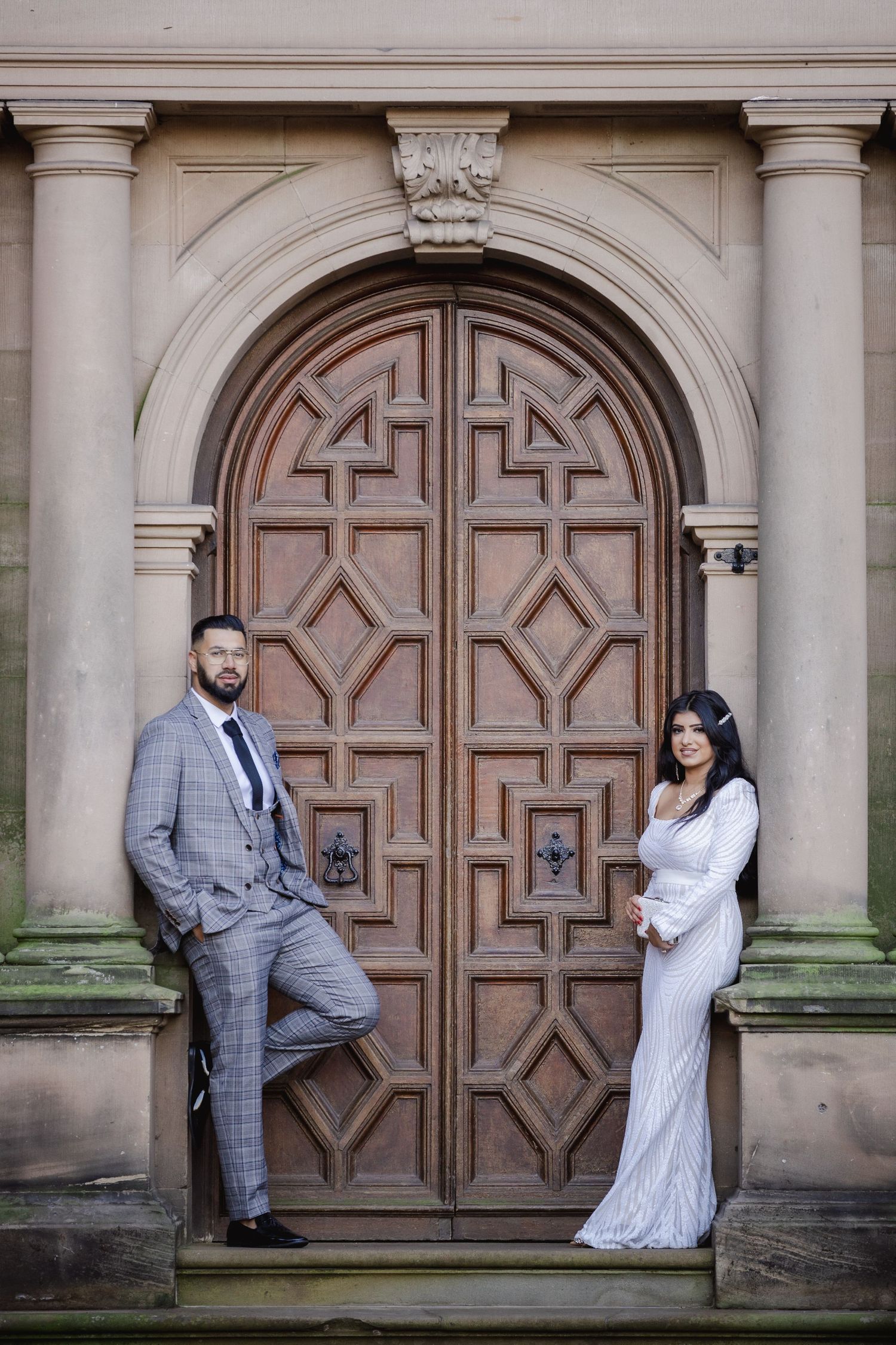 Couple in wedding attire posing before ornate wooden arched door of historic stone building.
