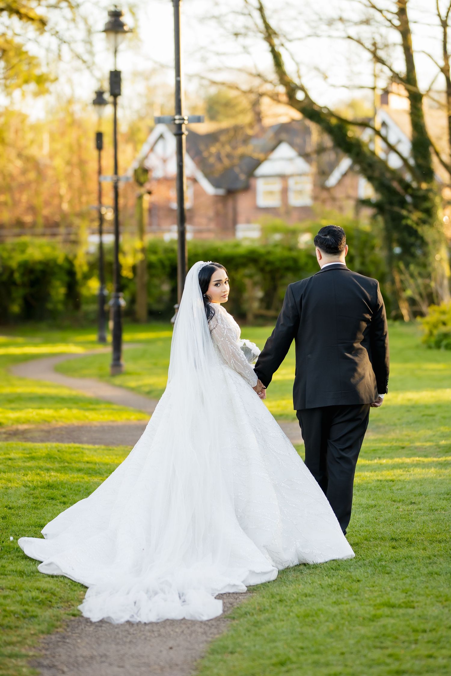 Bride in white gown and veil holds groom's hand walking through a lush green park on their wedding day.
