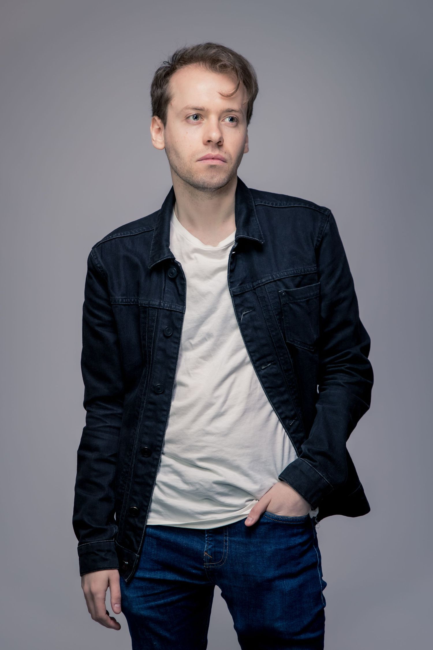 Young man in dark denim jacket and white t-shirt posing against gray background with hands in pockets.