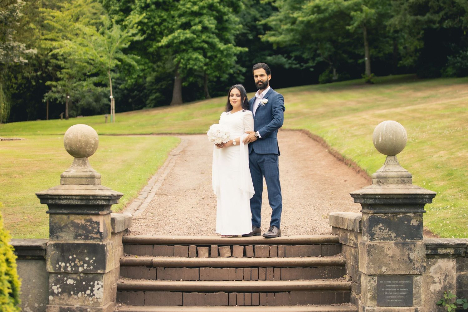 Bride and groom posing on stone steps in a lush garden setting, wearing white dress and navy suit.