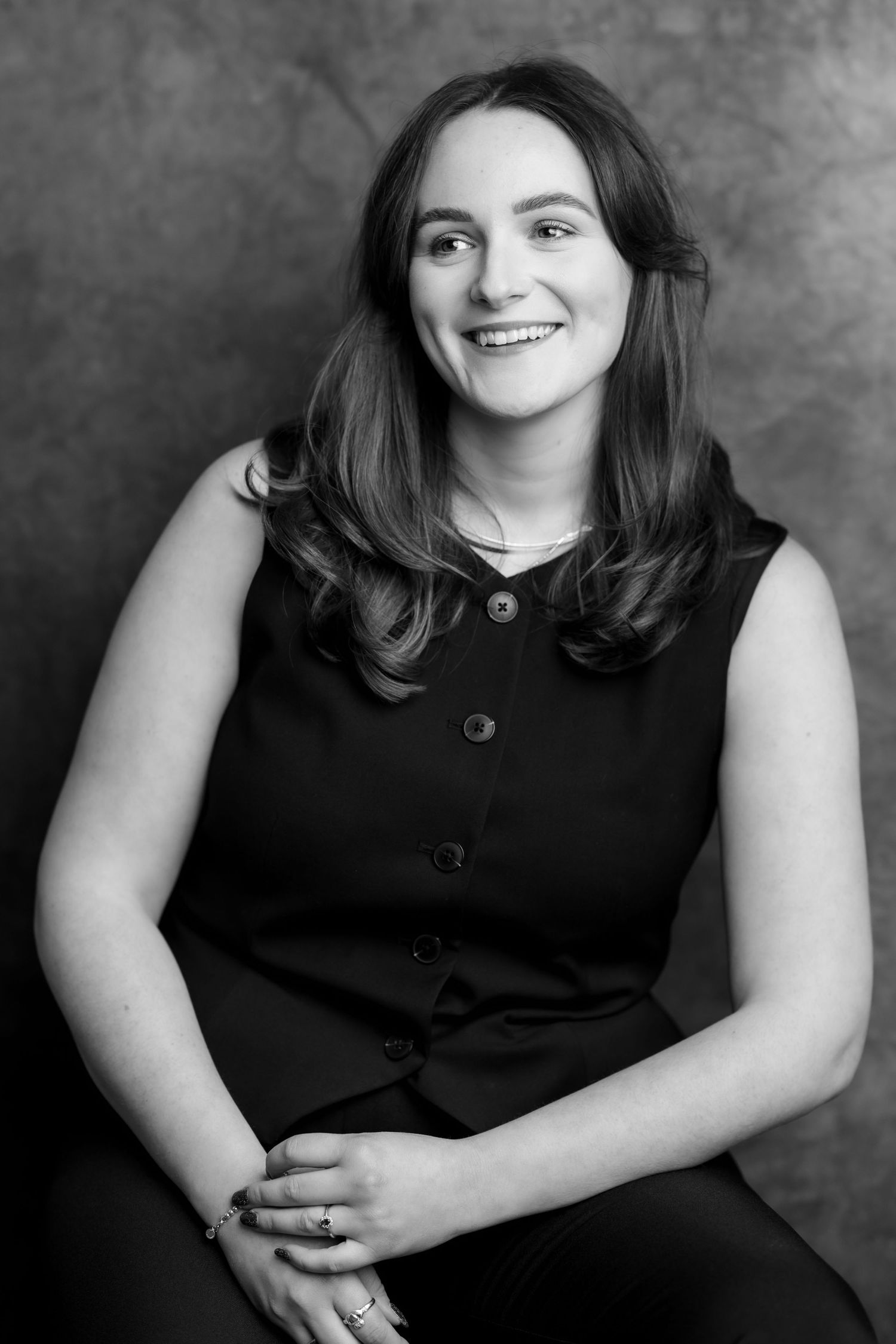 Black and white professional portrait of a smiling young woman in a sleeveless black top, seated with hands folded.