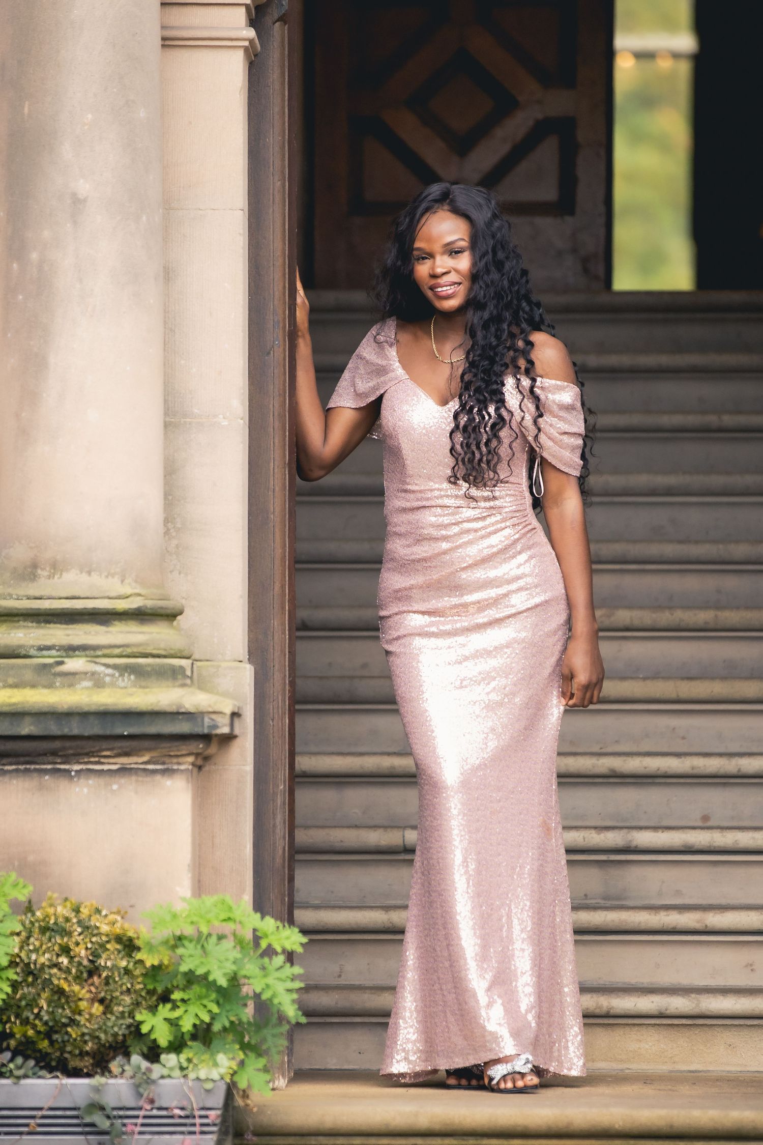 Black woman in pink sequin off-shoulder gown posing on stone steps of elegant building.