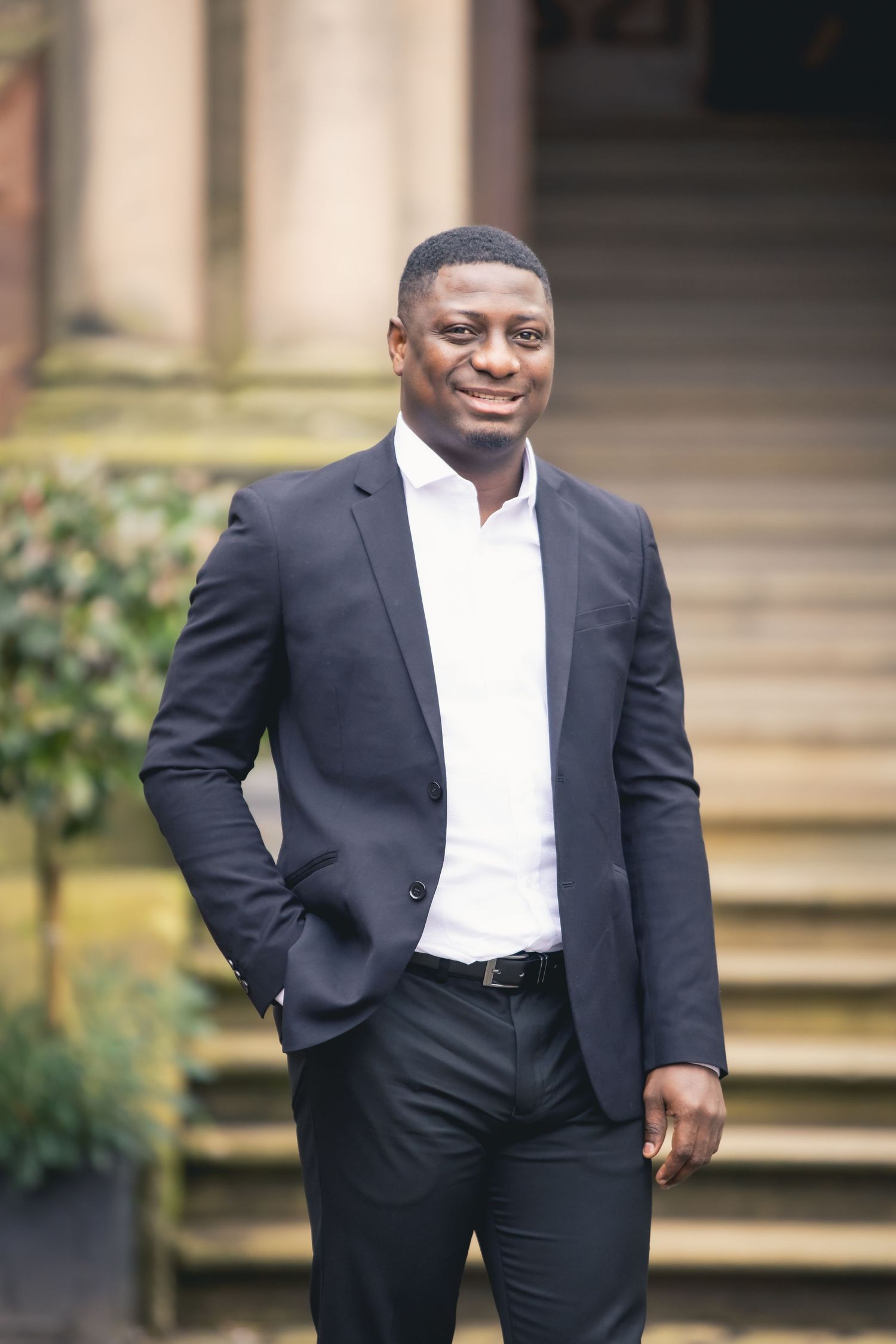 Smiling Black man in navy suit and white shirt standing outdoors near stone steps.