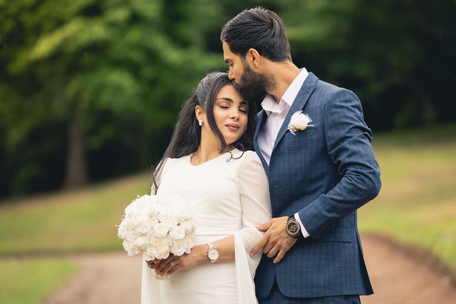 A groom in a blue suit kisses his bride wearing a white dress and holding a bouquet of white roses outdoors.