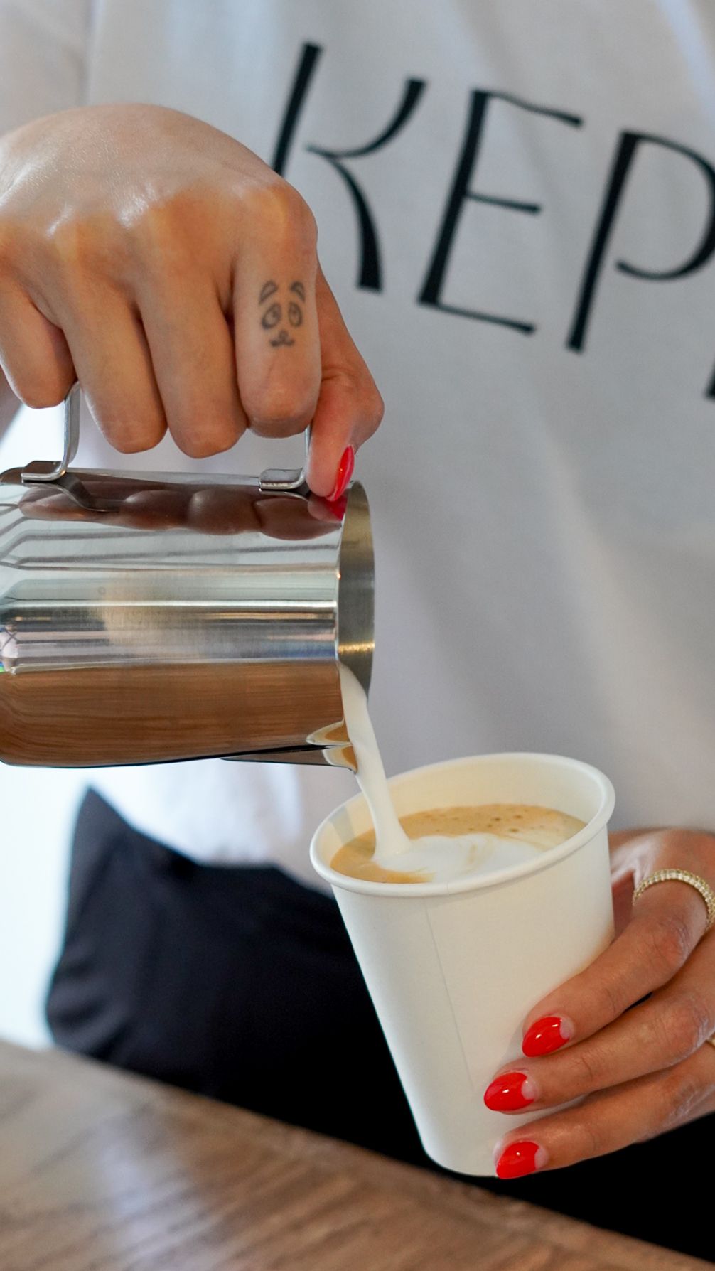 Barista with red nails and panda tattoo pouring steamed milk from steel pitcher into white paper cup.