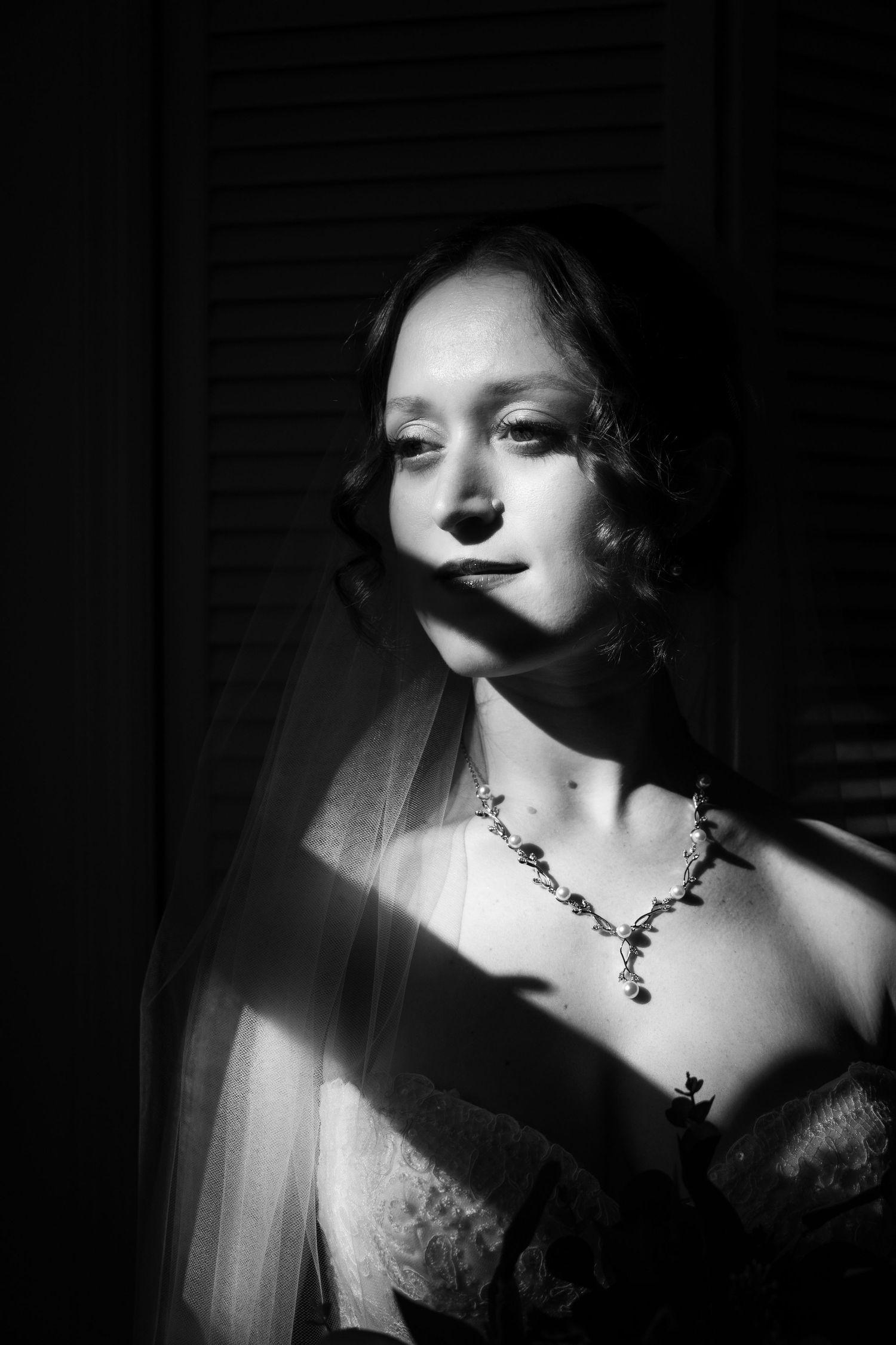 Black and white portrait of a bride in veil and floral necklace, dramatically lit with shadows.