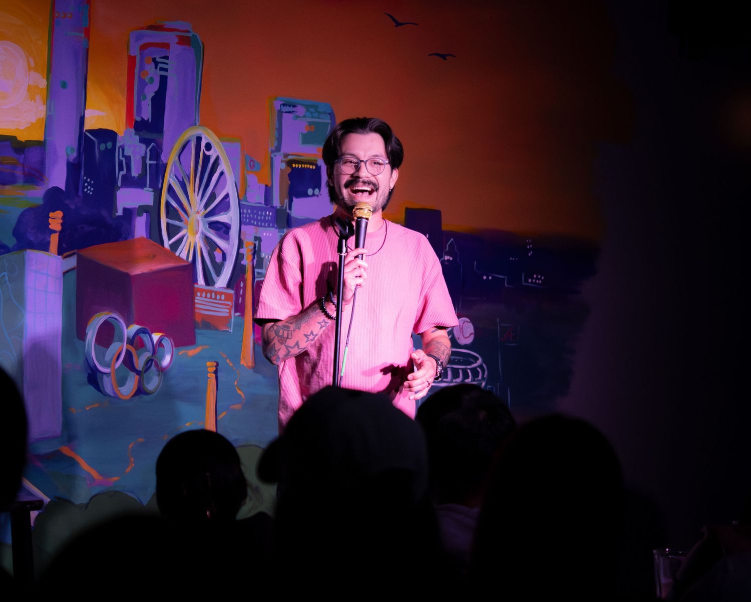 A comedian performs stand-up on a colorful stage, smiling into a microphone wearing a pink shirt.