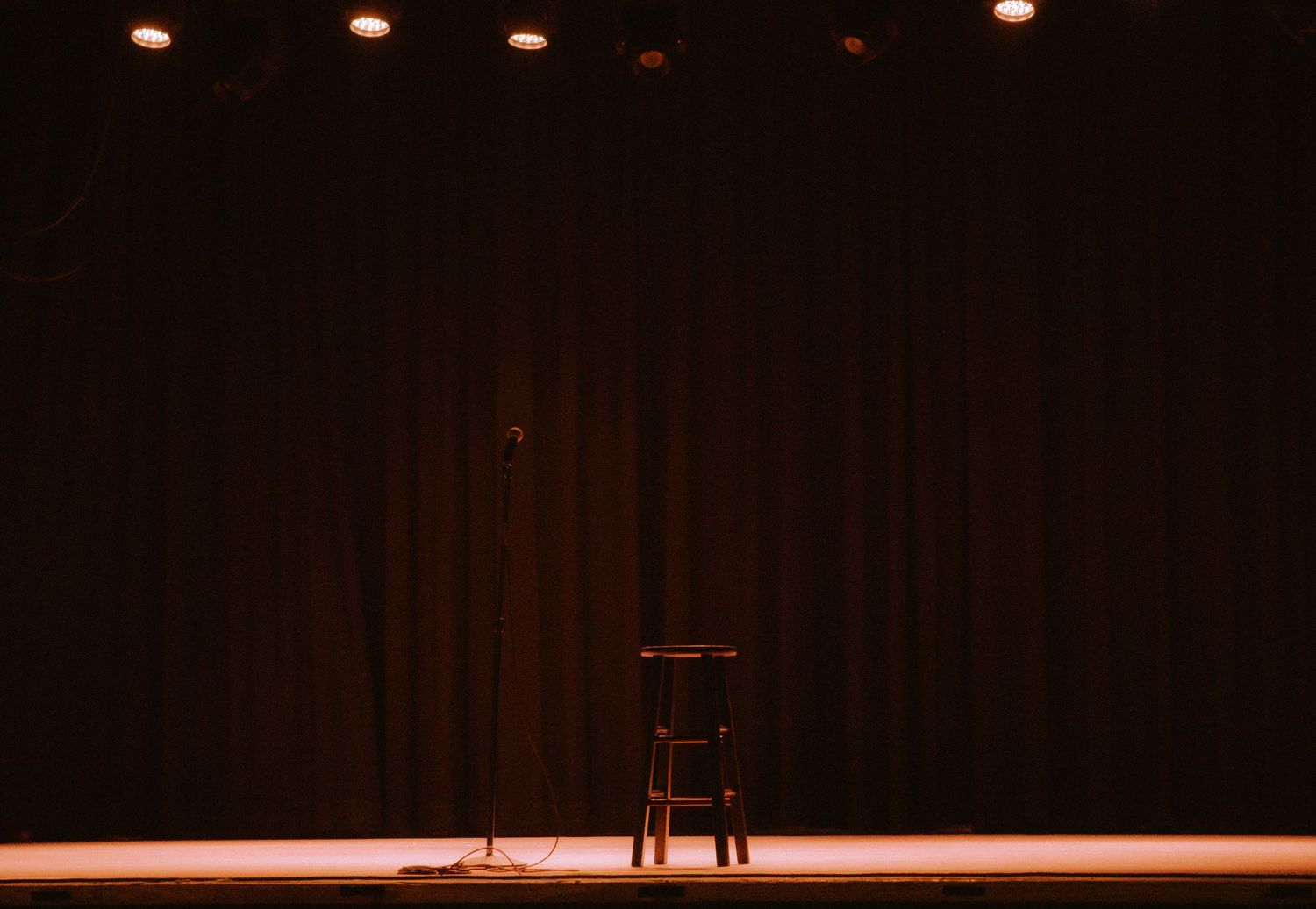 Empty comedy club stage with microphone stand, stool, and dramatic spotlight against dark curtain backdrop.